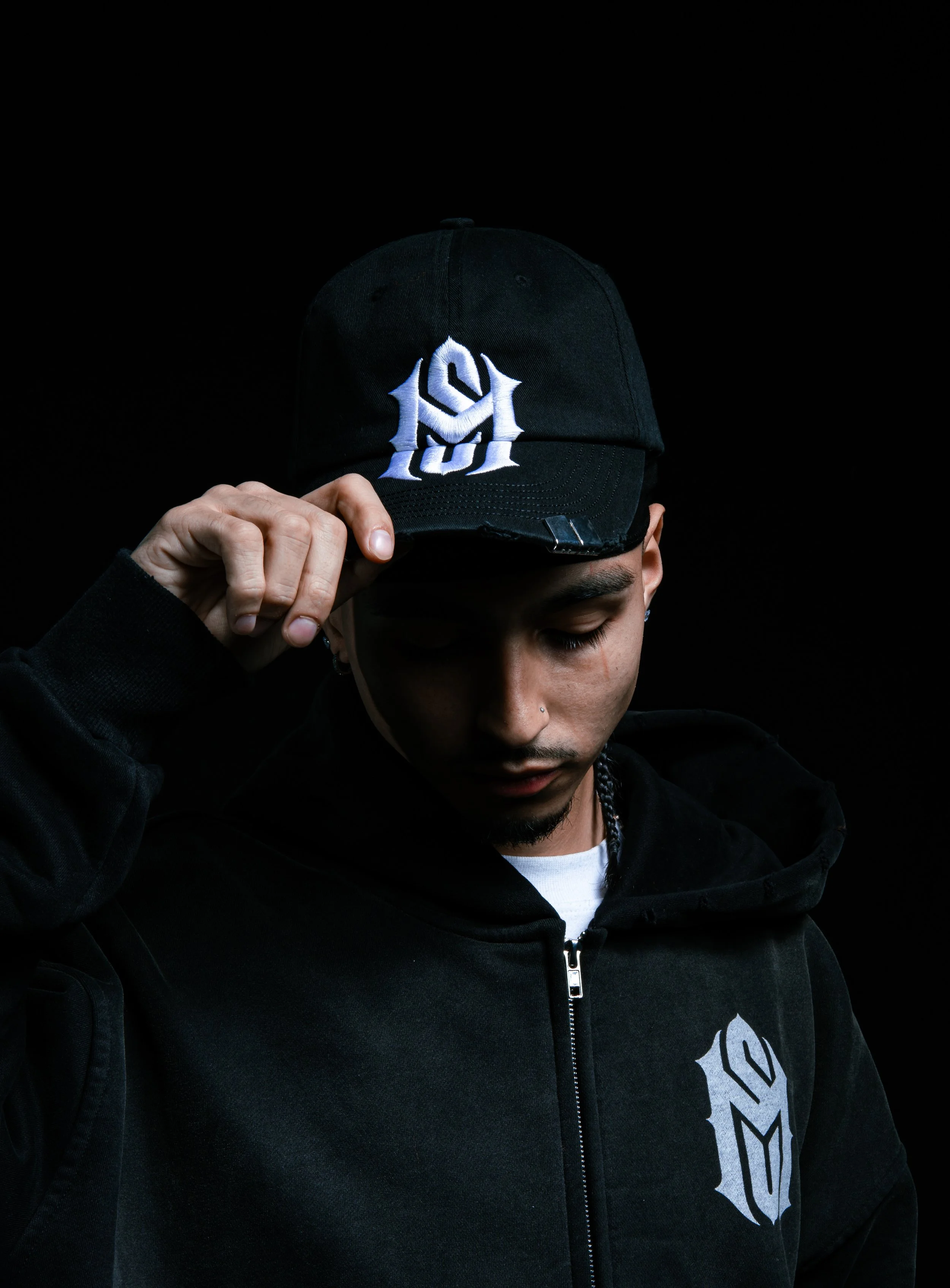 Young man wearing a black hoodie and a black cap with a white logo, against a black background.