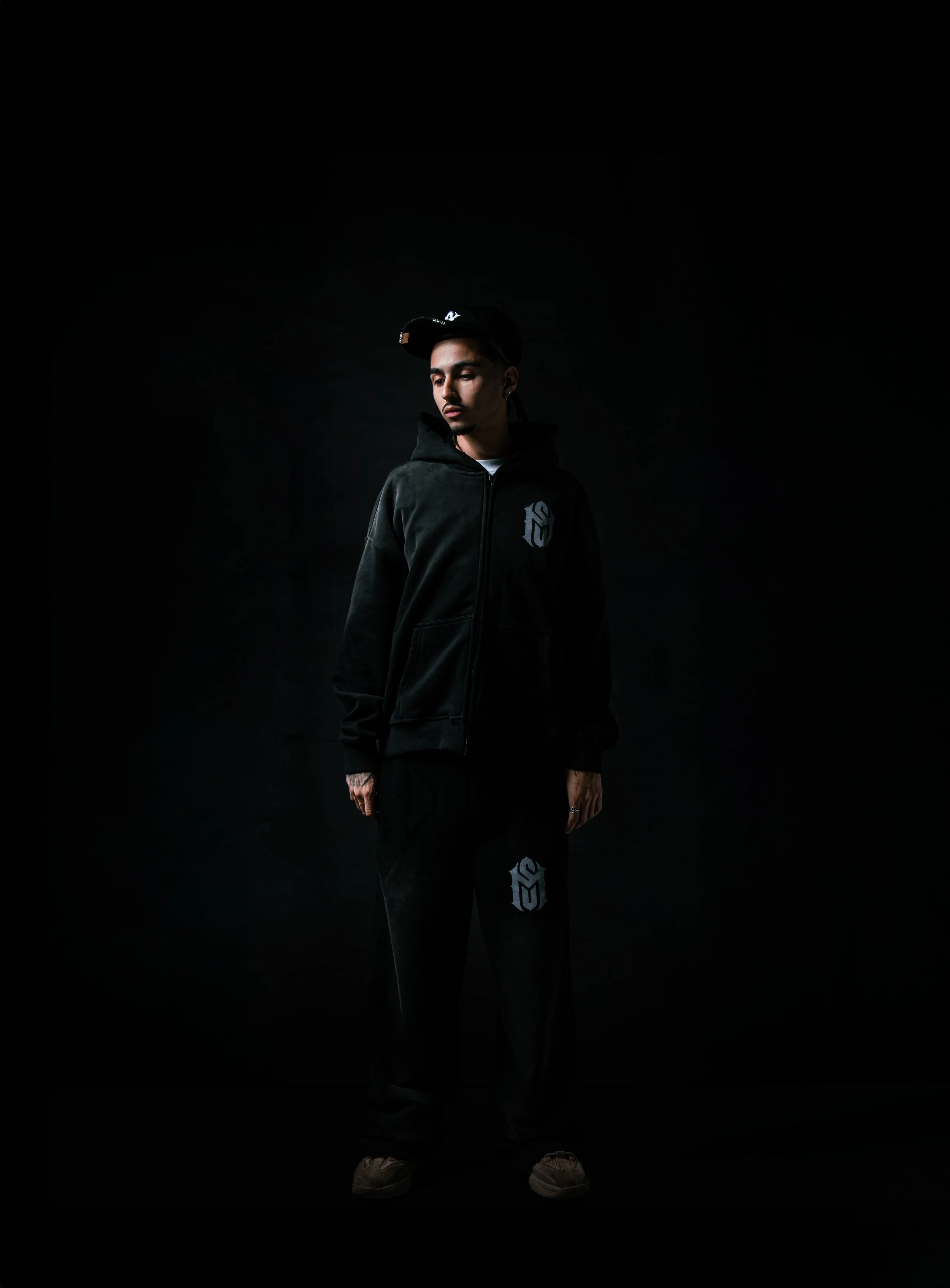 Young man in black hoodie and sweatpants standing against a dark background, with a black cap, looking to the side.