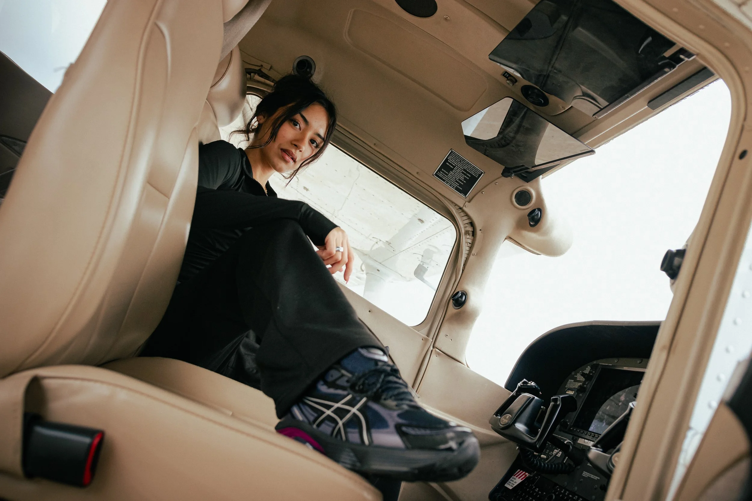 A woman sitting inside the cockpit of a helicopter, looking towards the camera. She is dressed in black and wearing sneakers.