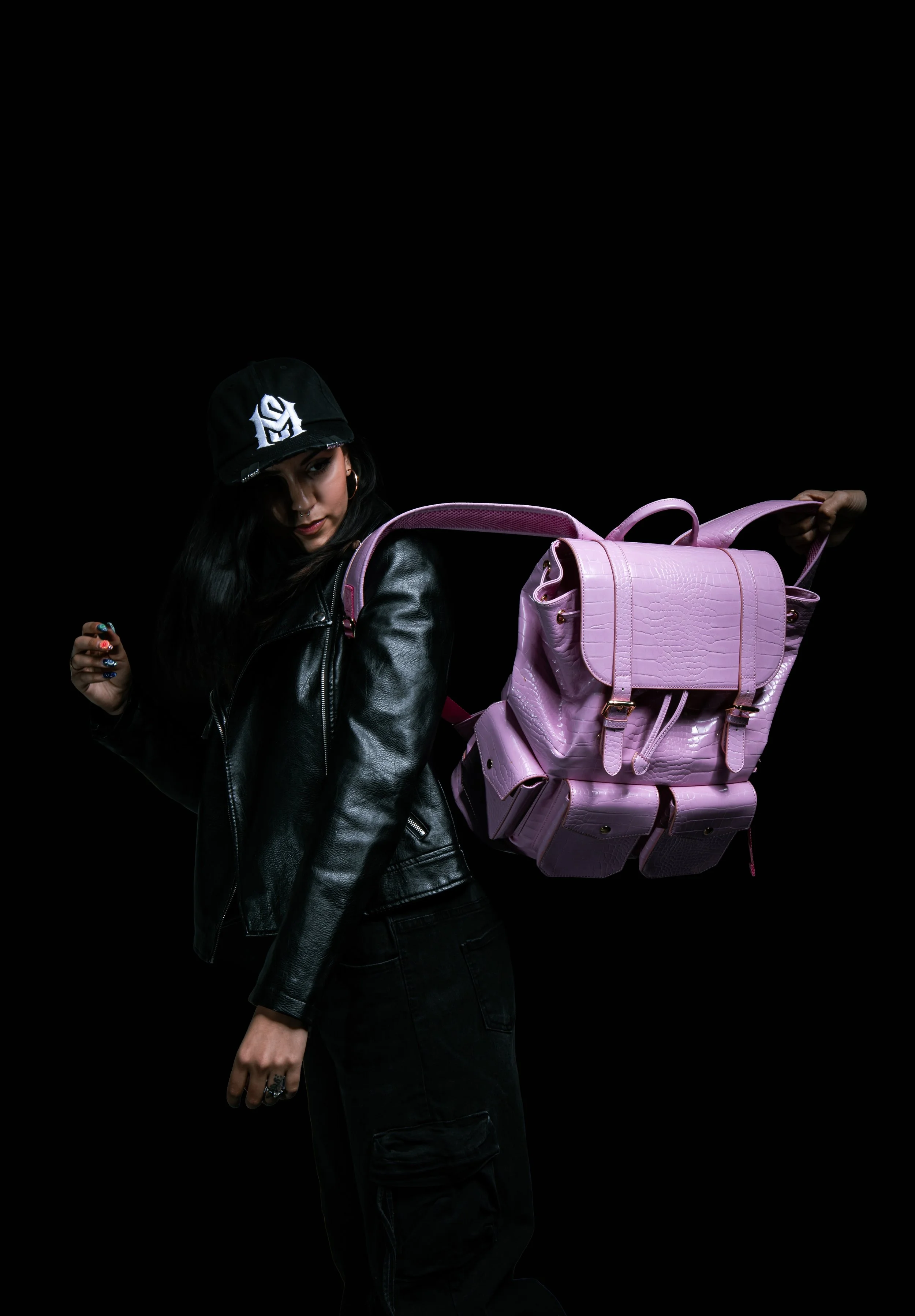 A woman with long black hair wearing a black leather jacket, a black cap, and hoop earrings, holding a large pink backpack with crocodile texture against a black background.