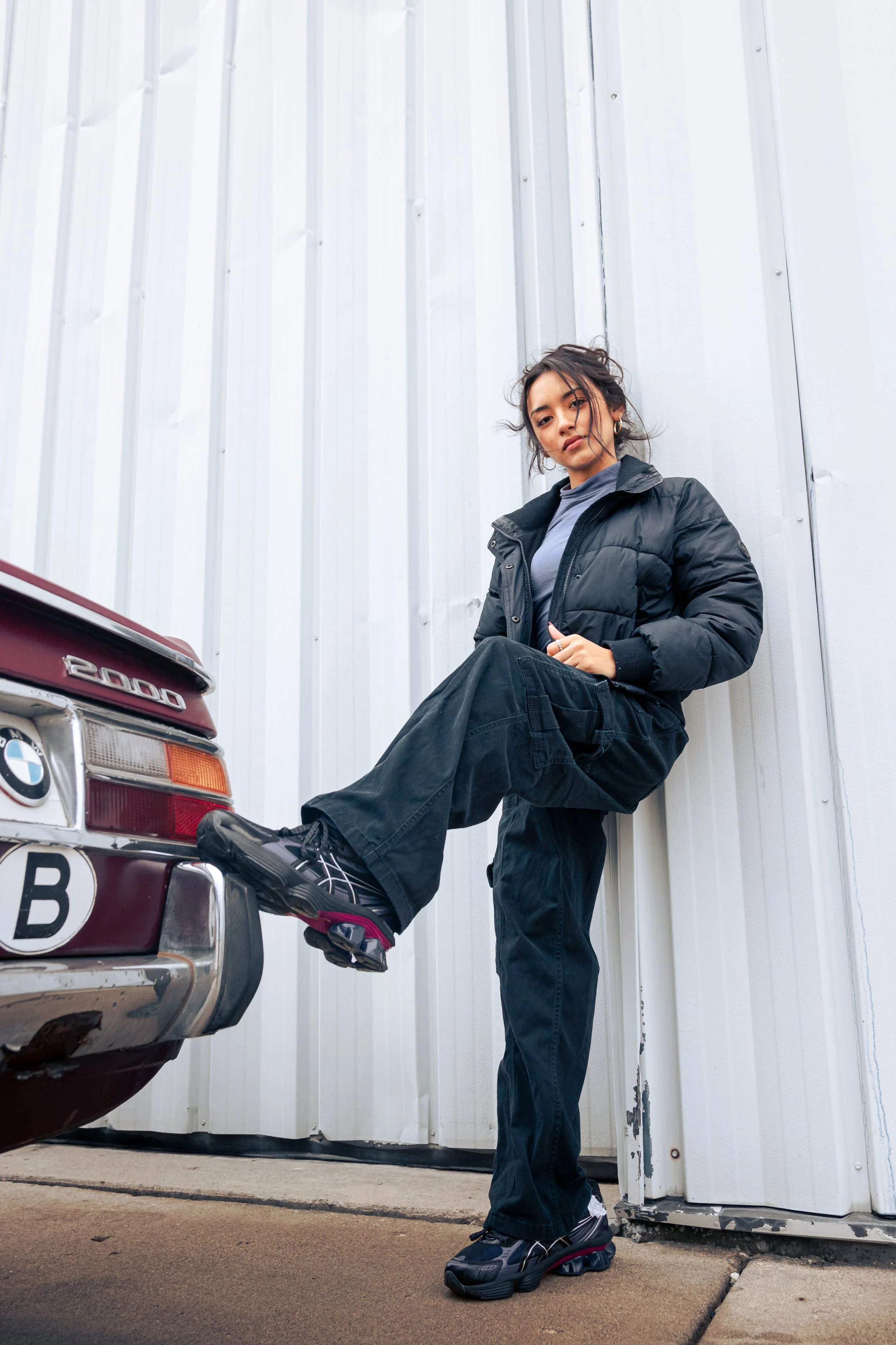 Young woman in black puffer jacket and black pants leaning against a white wall, with one foot resting on the rear of a vintage BMW car.