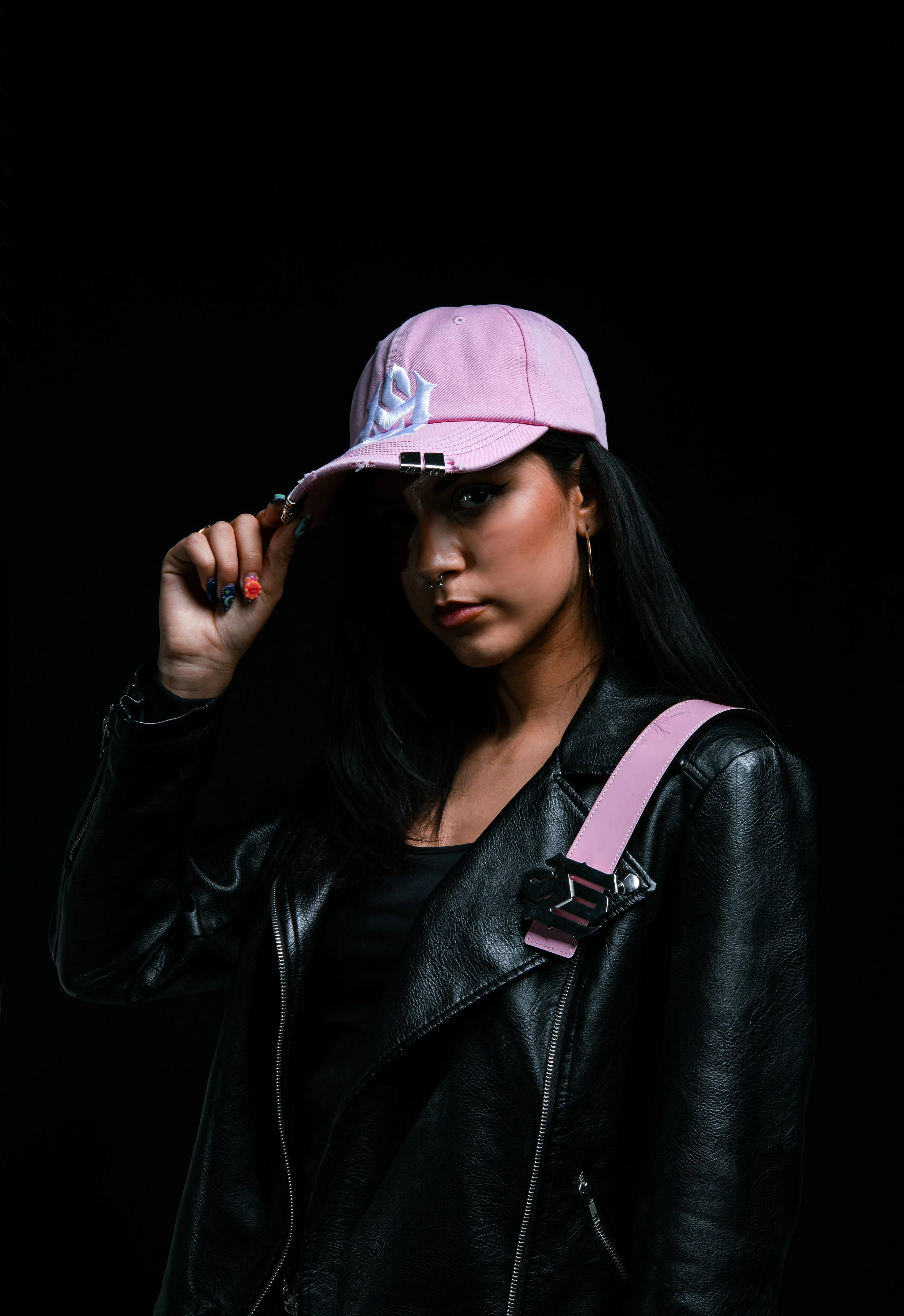 A young woman with black hair wearing a black leather jacket, hoop earrings, and a pink baseball cap, posing against a black background.