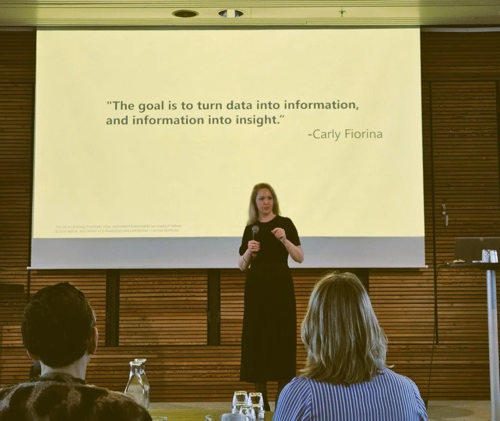 Dr.Kolset in a black dress is speaking at a presentation with a microphone in front of a large screen displaying a quote by Carly Fiorina about turning data into insight. The audience is seated, facing the speaker and the screen.