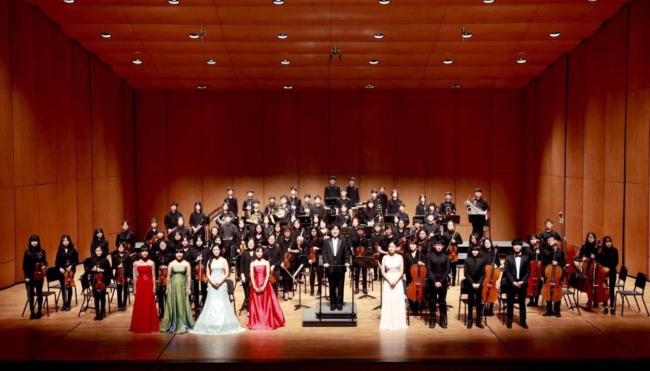 An orchestra standing on stage after a performance, with musicians in black attire and a few in formal dresses, in a concert hall with wood-paneled walls.