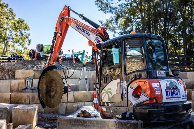 Sunshine Coast Sandstone image of Mini excavator with a saw attachment working on a sandstone retaining wall during construction or landscaping.