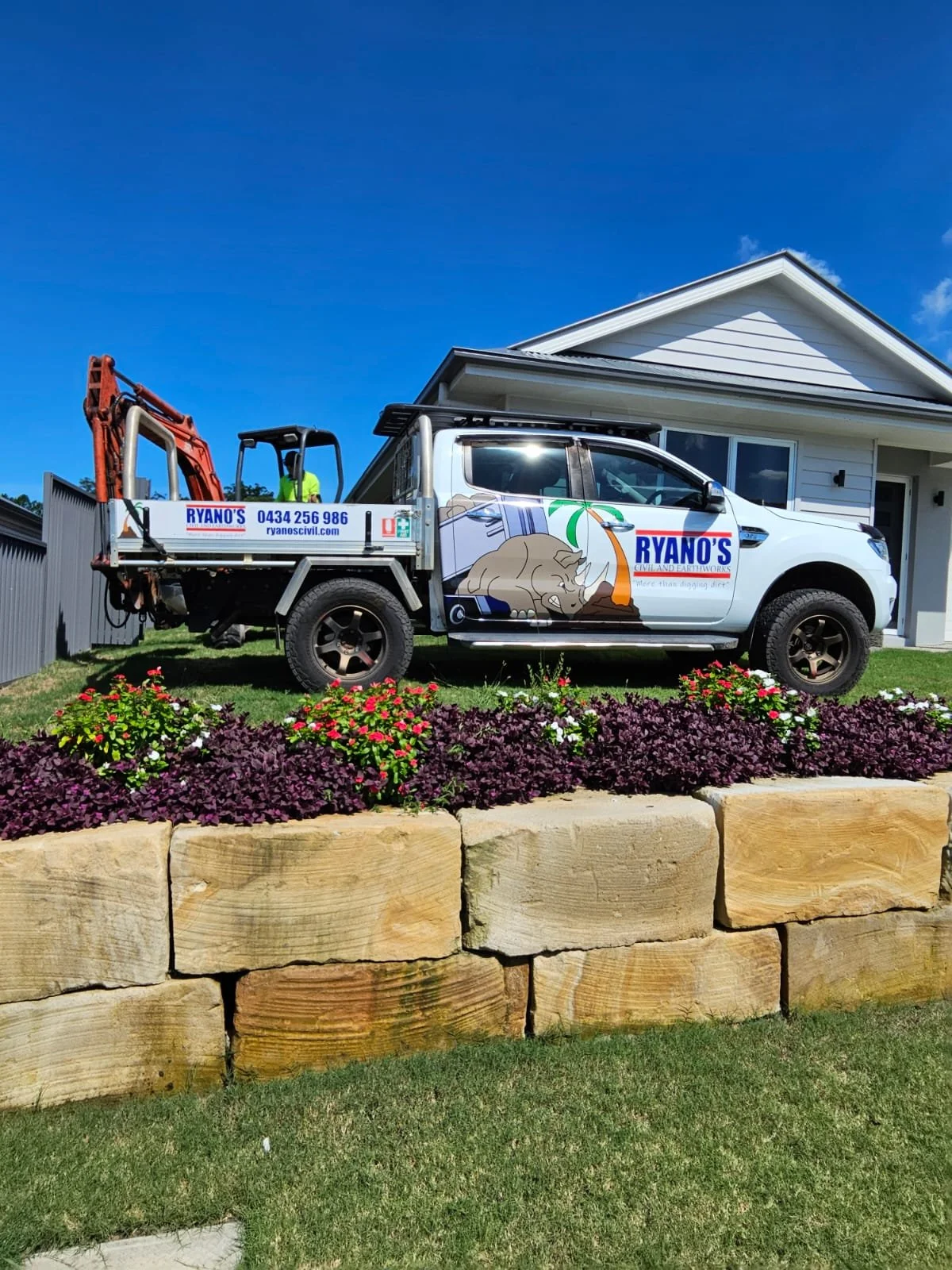 White service vehicle with Ryano's Civil And Earthworks branding, parked on a manicured lawn in front of a house, with flowers and sandstone block retaining.