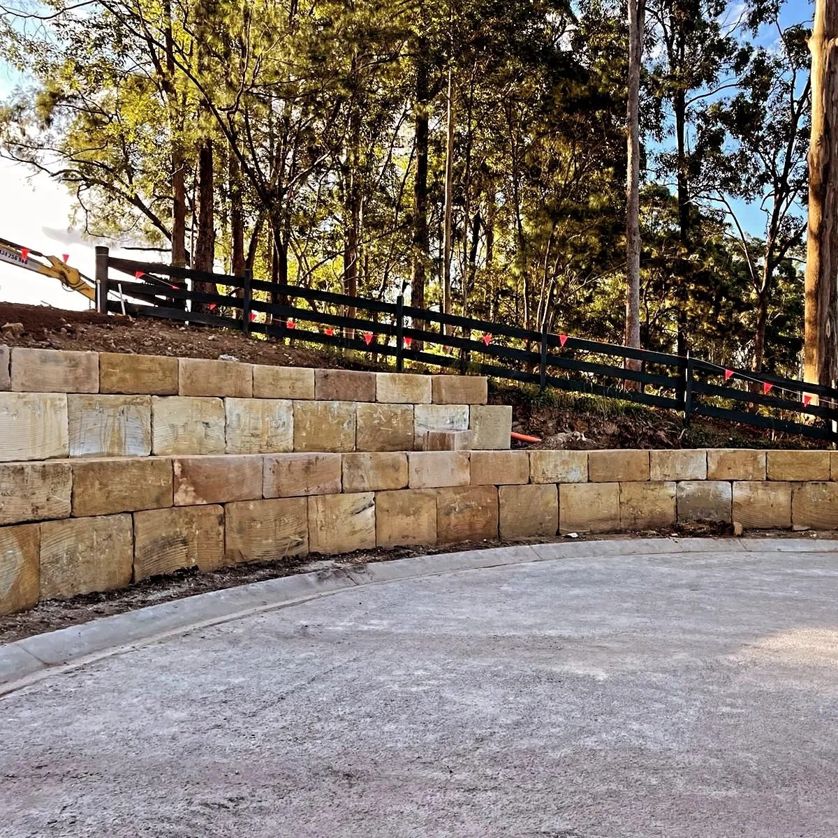 Sunshine Coast Sandstone image of a curved concrete driveway bordered by a sandstone retaining wall and a black wooden fence, with a hillside of trees in the background under a partly cloudy sky.