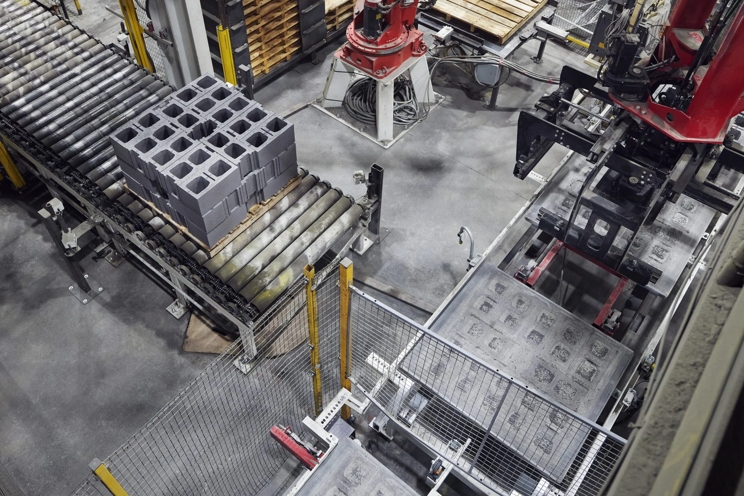Industrial factory floor with conveyor belt carrying molded concrete blocks, robotic arms, and machinery for manufacturing or processing materials.