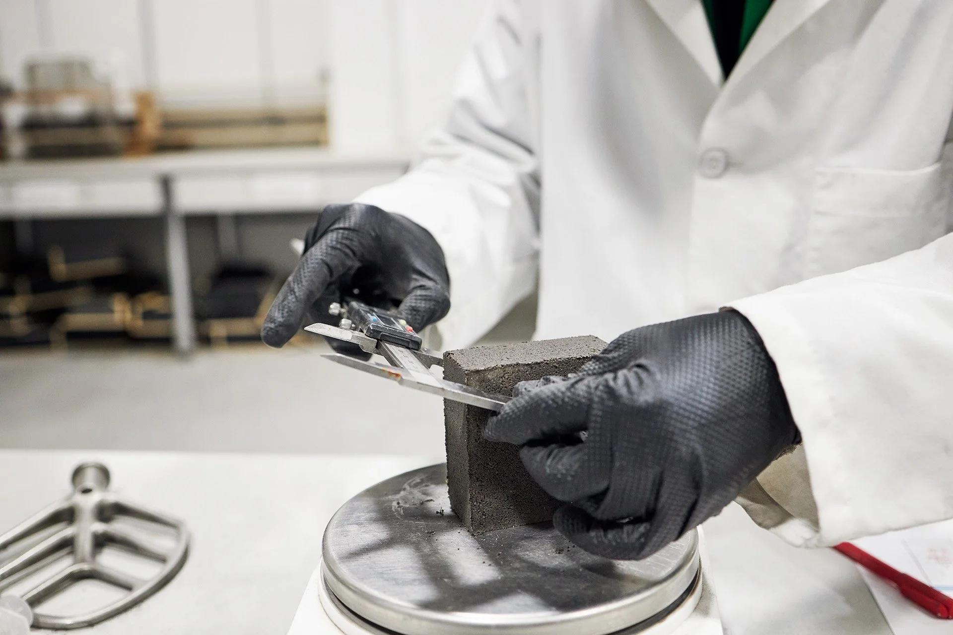 Person wearing black gloves and a white lab coat holding a mortar sample on a scale in a laboratory setting.