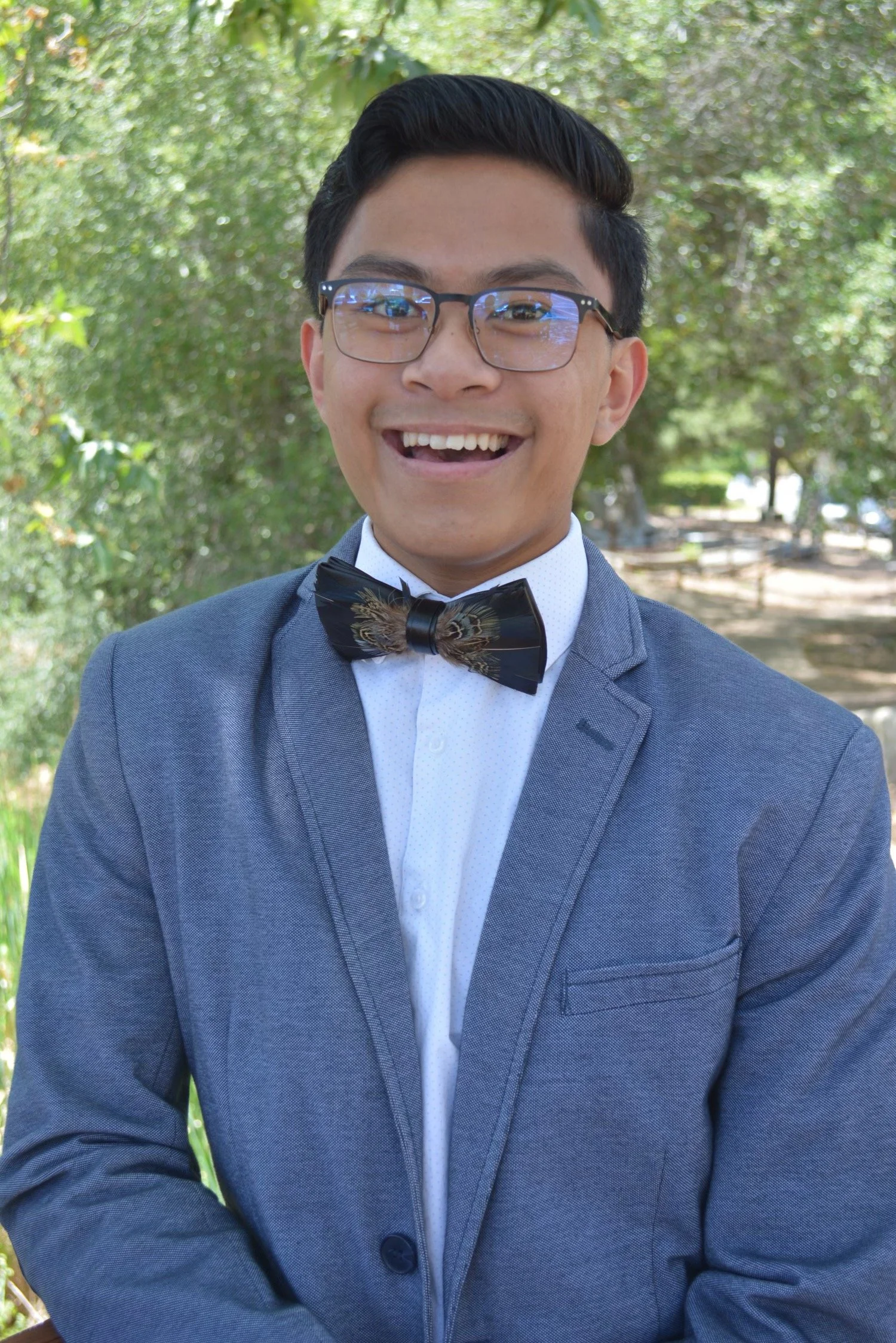 A young man wearing glasses, a gray suit, a white shirt, and a bow tie, smiling outdoors with trees and greenery in the background.