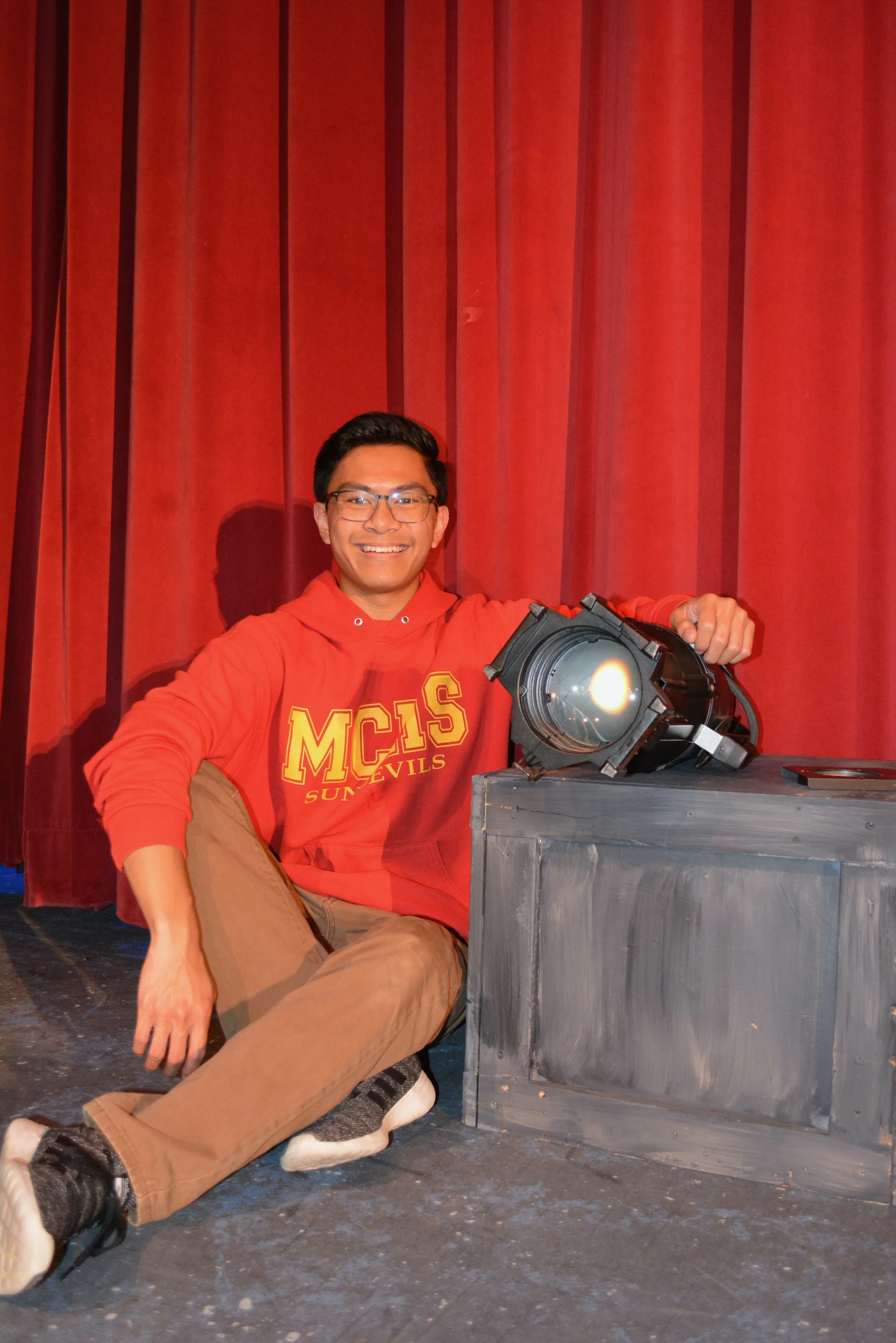 A smiling young man sitting on the floor in front of red stage curtains, wearing glasses, a red MCIS Sun Devils hoodie, and tan pants, holding a large stage light.