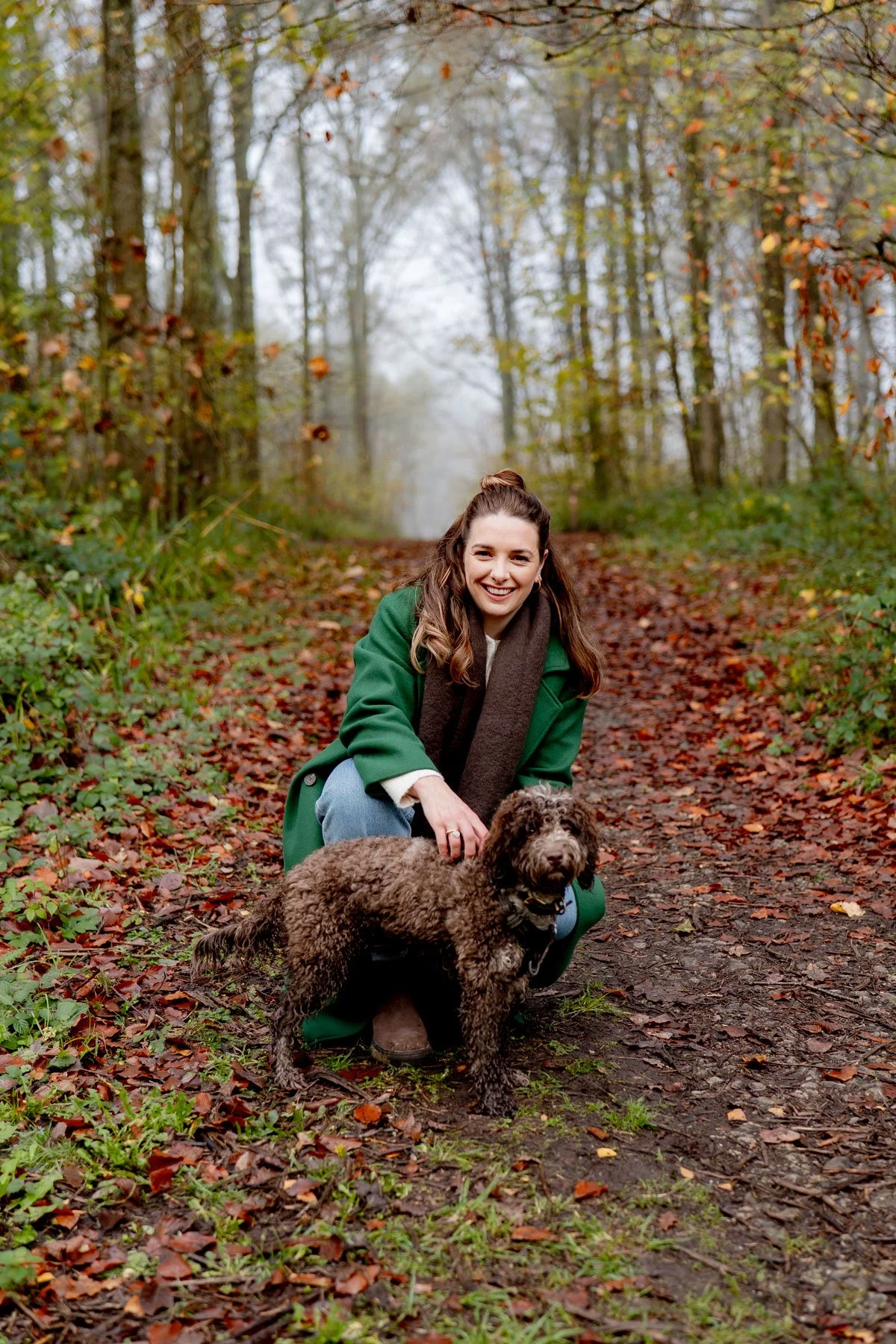 A woman smiling and crouching on a wooded trail with a curly-haired dog during autumn.