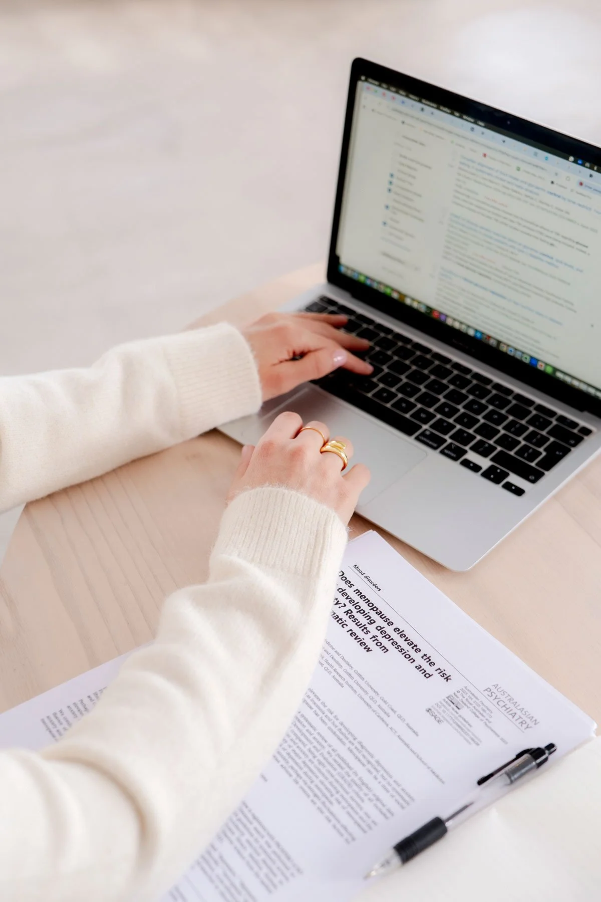 Person in a white sweater working on a laptop with a document and pen on a light-colored table.