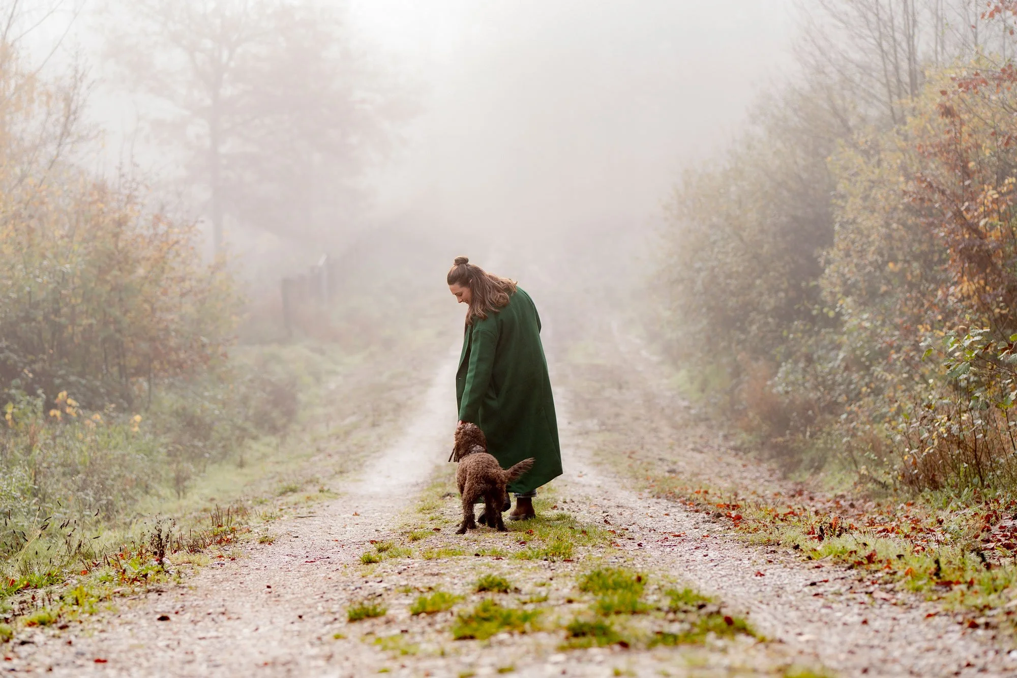 A woman in a green coat walking her dog on a foggy dirt path surrounded by autumn trees.