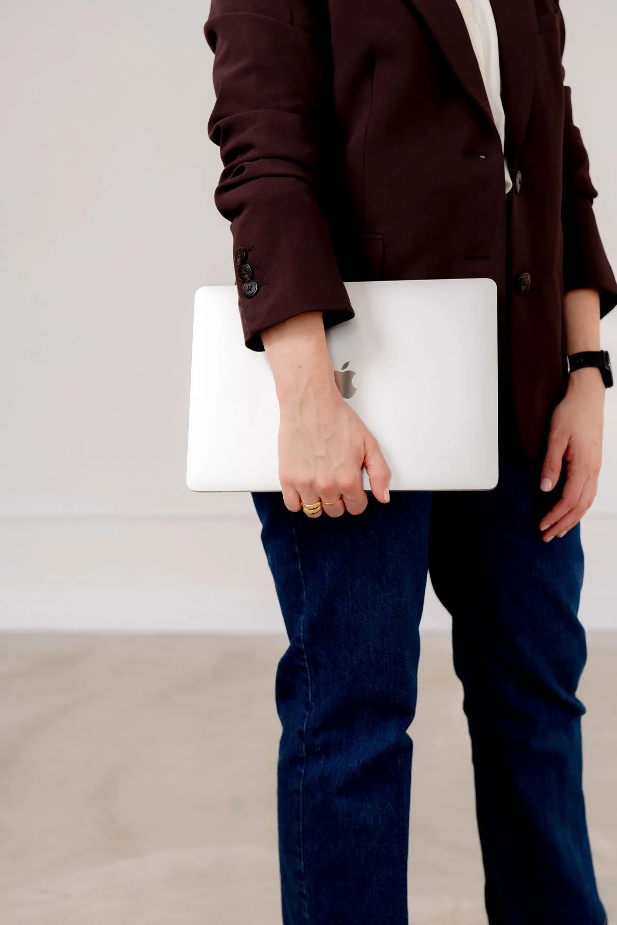 Person holding a silver MacBook with Apple logo, wearing a dark red blazer, blue jeans, and a black watch, standing indoors against a plain light background.