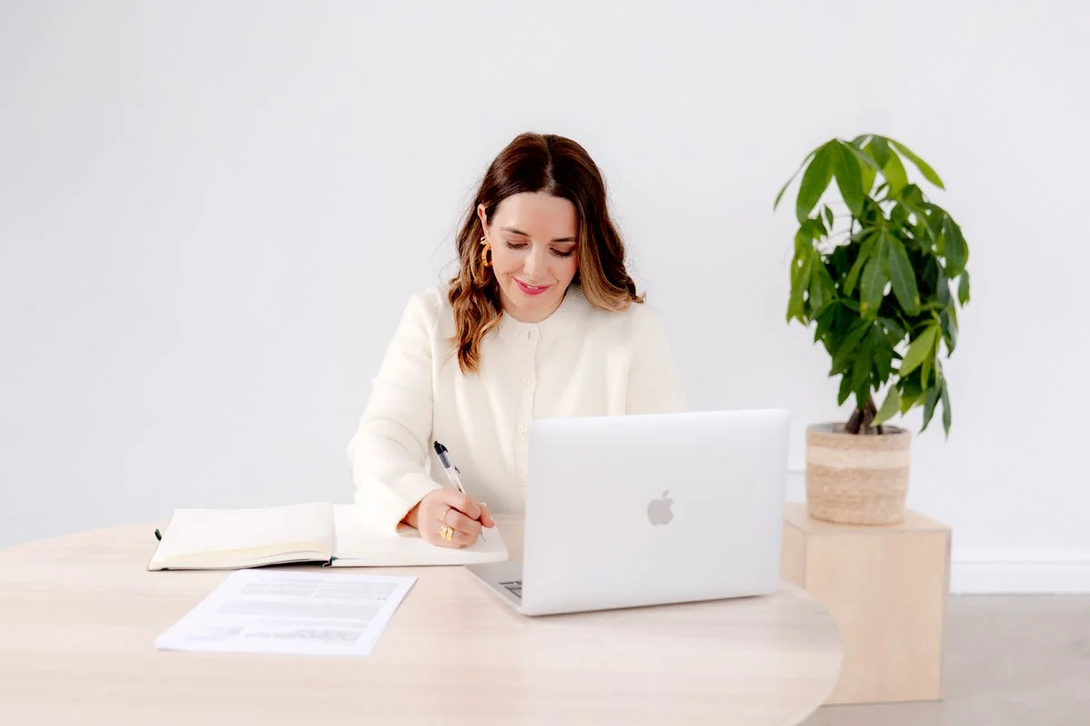 A woman sitting at a desk with a silver laptop, notebook, pen, and papers in front of her, smiling and writing, with a potted green plant on her right side against a white background.