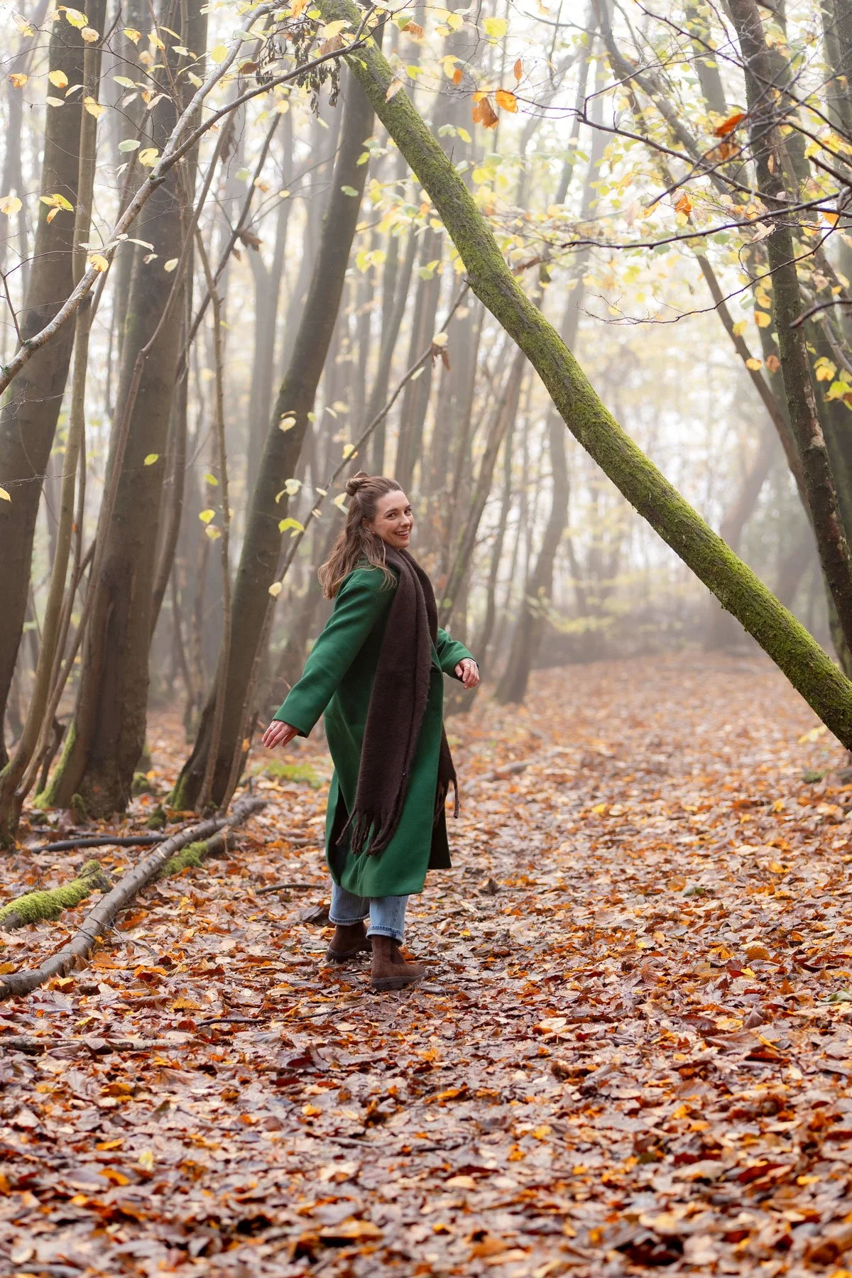 A woman with long brown hair in a bun, wearing a green coat, brown scarf, blue jeans, and brown boots, smiling and walking through an autumn forest covered with fallen leaves.