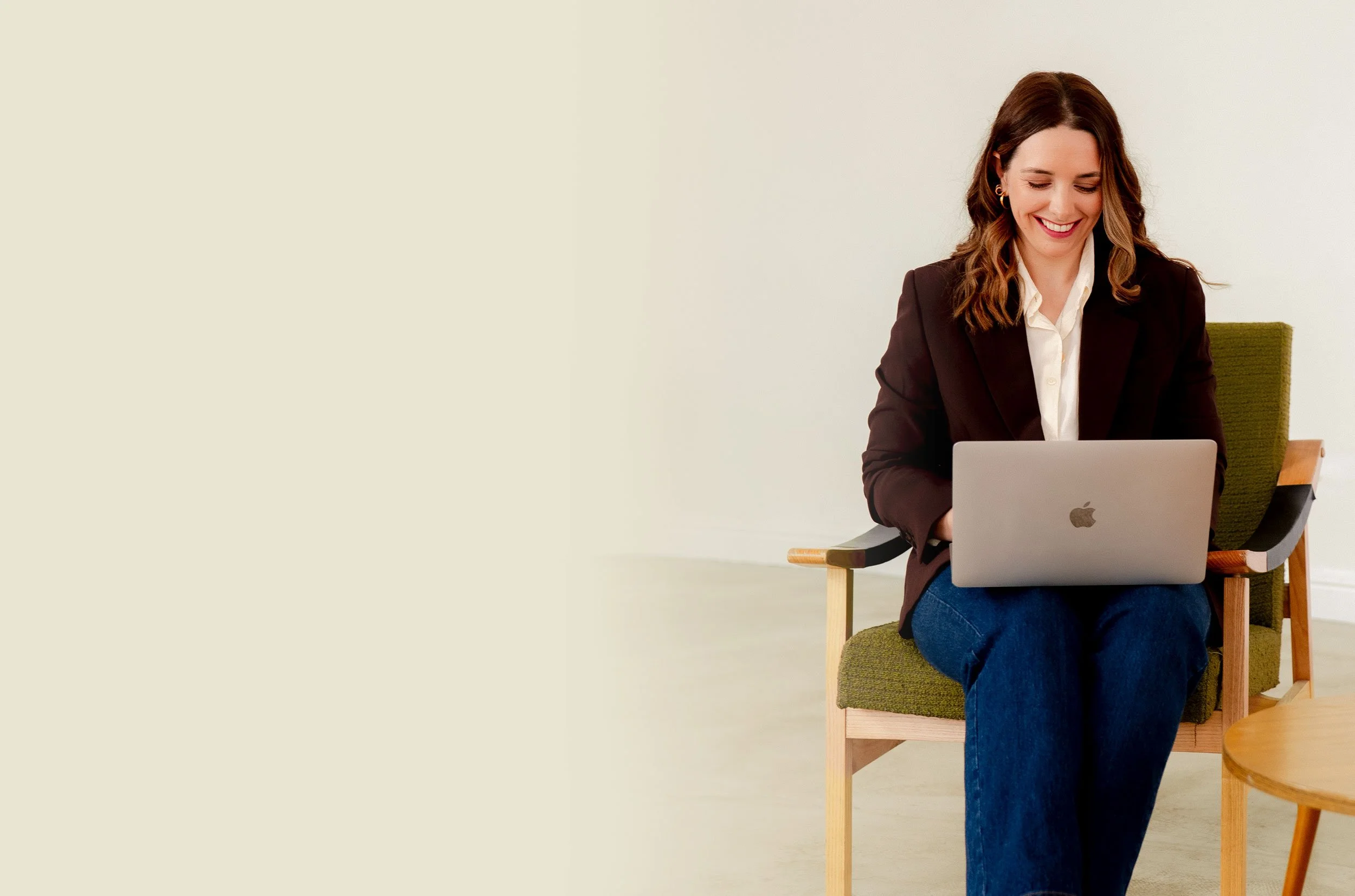 A woman sitting on a green chair with wooden arms, using a silver MacBook, smiling, dressed in a dark blazer, white shirt, and blue jeans.