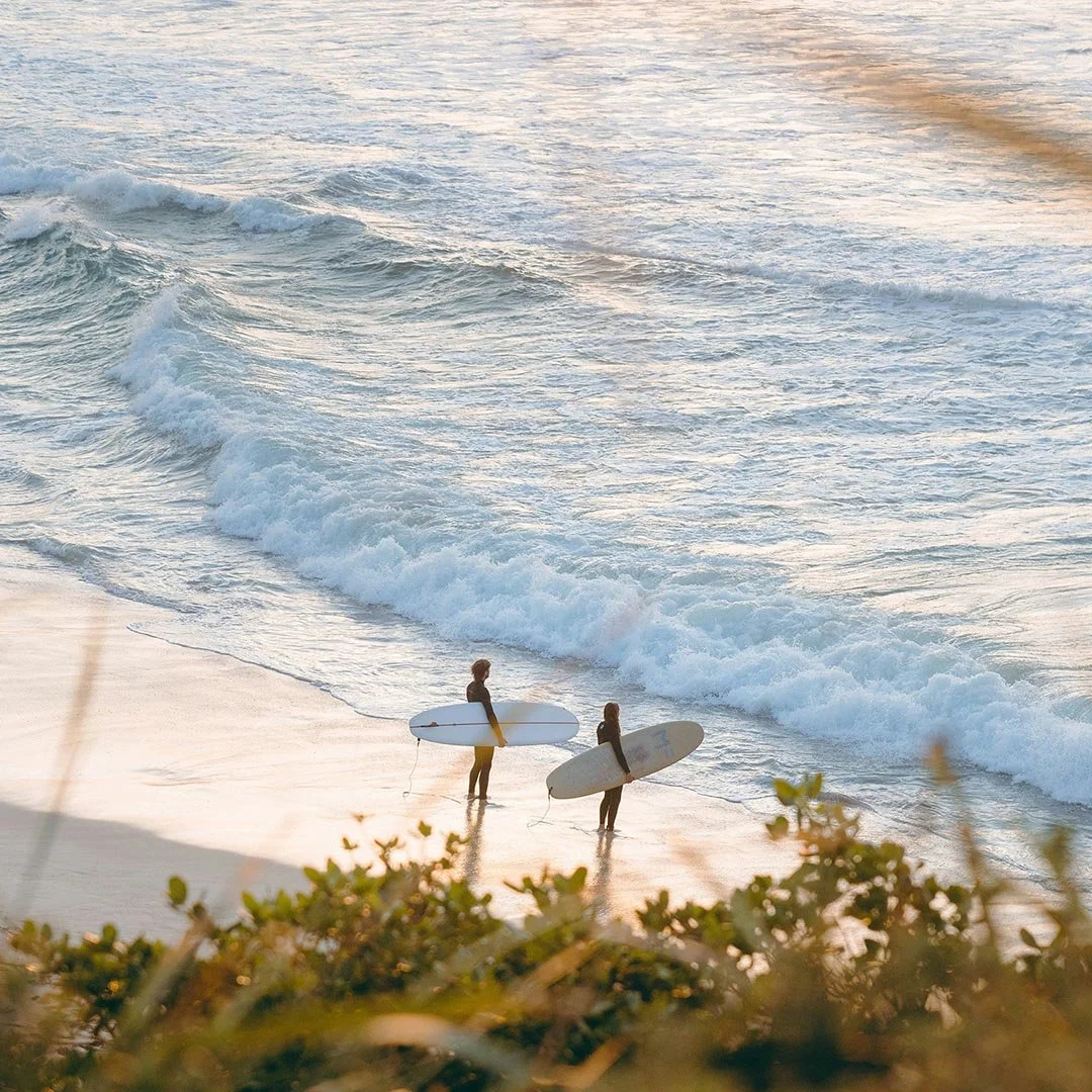 Two surfers, one man and one woman, standing on the sandy beach holding surfboards, facing the ocean with waves approaching during sunset on the Mornington Peninsula.