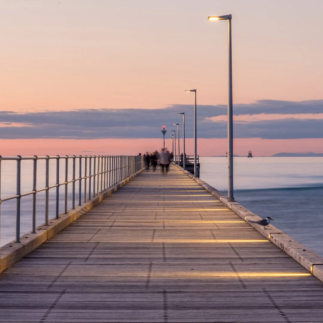 The Rosebud Pier at sunset, with streetlights and a few people walking. The sky is pink and blue with clouds, and there are distant islands and an oil platform visible in the water. Rosebud, Mornington Peninsula.