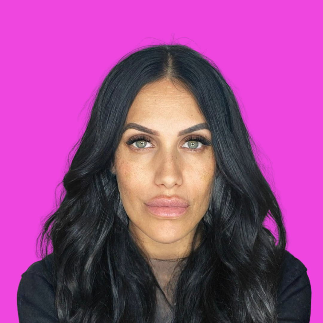 Close-up portrait of a woman with long black hair, green eyes, and makeup, set against a vivid pink background.
