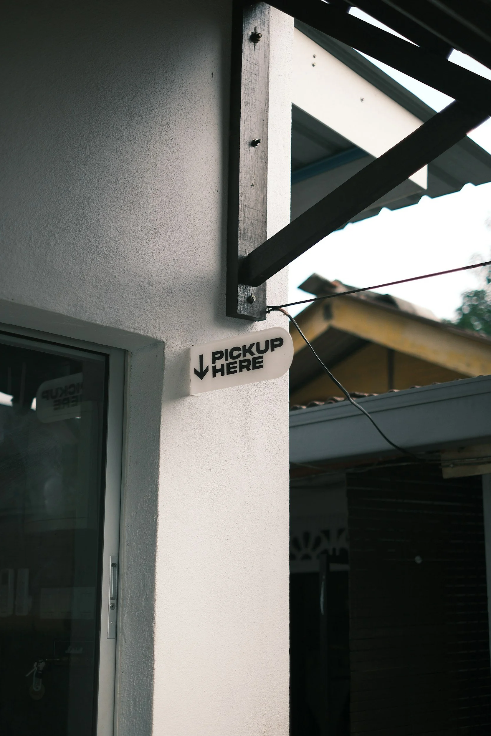 A small white sign with black text and an arrow pointing downward reading 'PICKUP HERE' hangs outside a building near a glass door, under a wooden structure.