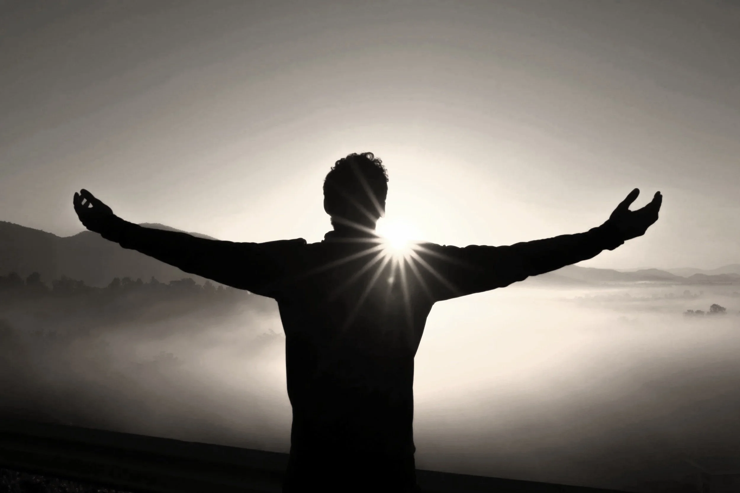 Silhouette of a person with arms outstretched standing outside during sunrise or sunset, with sun rays shining behind them and a foggy landscape in the background.