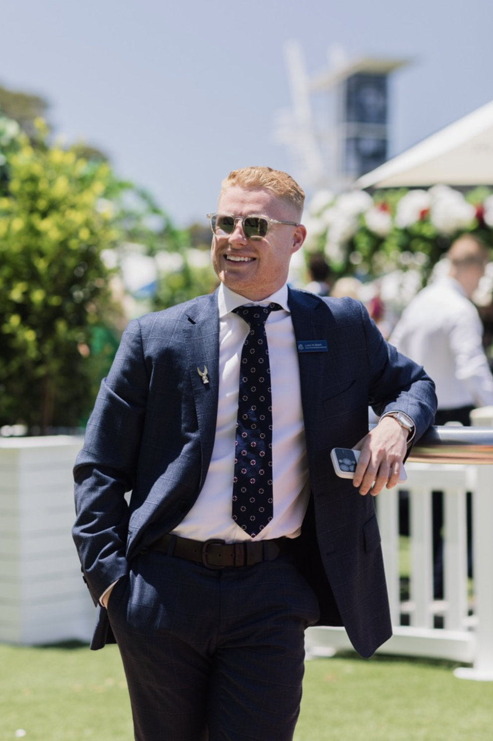 Man in a navy checkered suit, white shirt, and patterned tie, wearing sunglasses, outdoors at an event, smiling, holding a phone in his left hand, with greenery and other people in the background.
