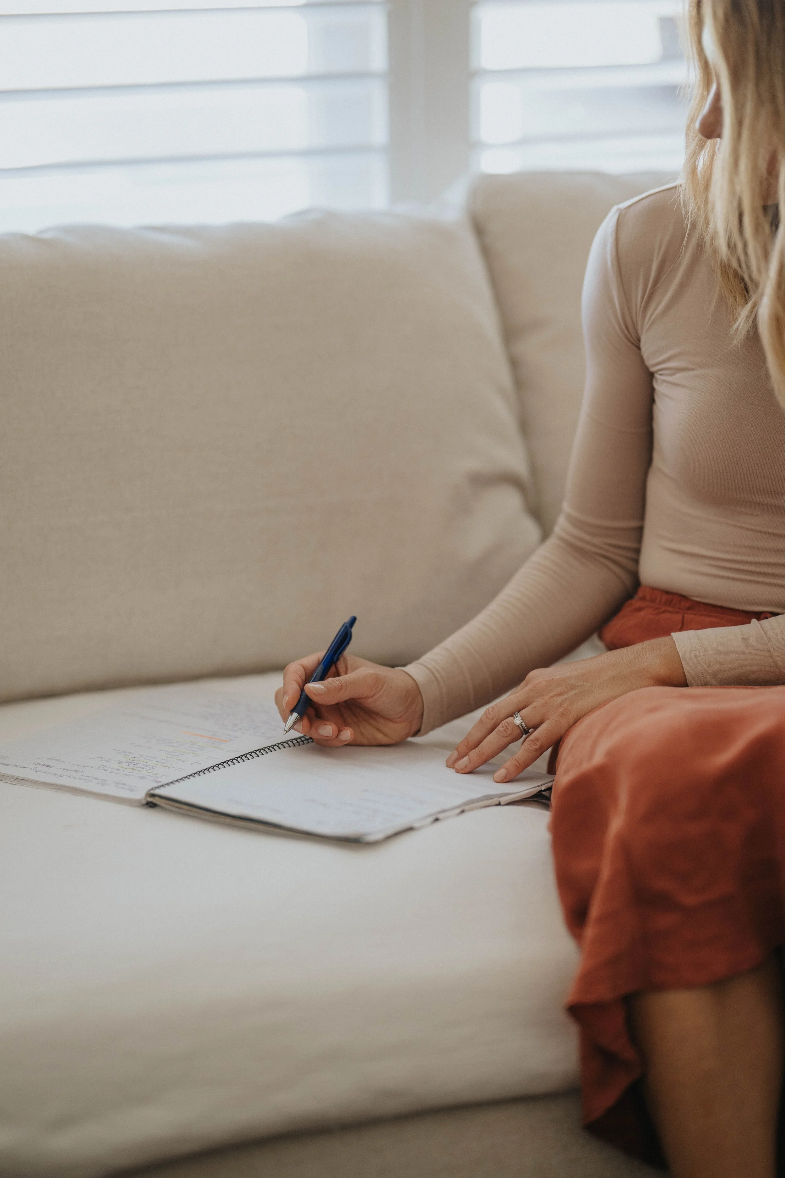 A woman sitting on a cream-colored sofa, writing in a notebook with a blue pen. She is wearing a beige long-sleeve shirt and a rust-colored skirt. The background features a window with blinds.
