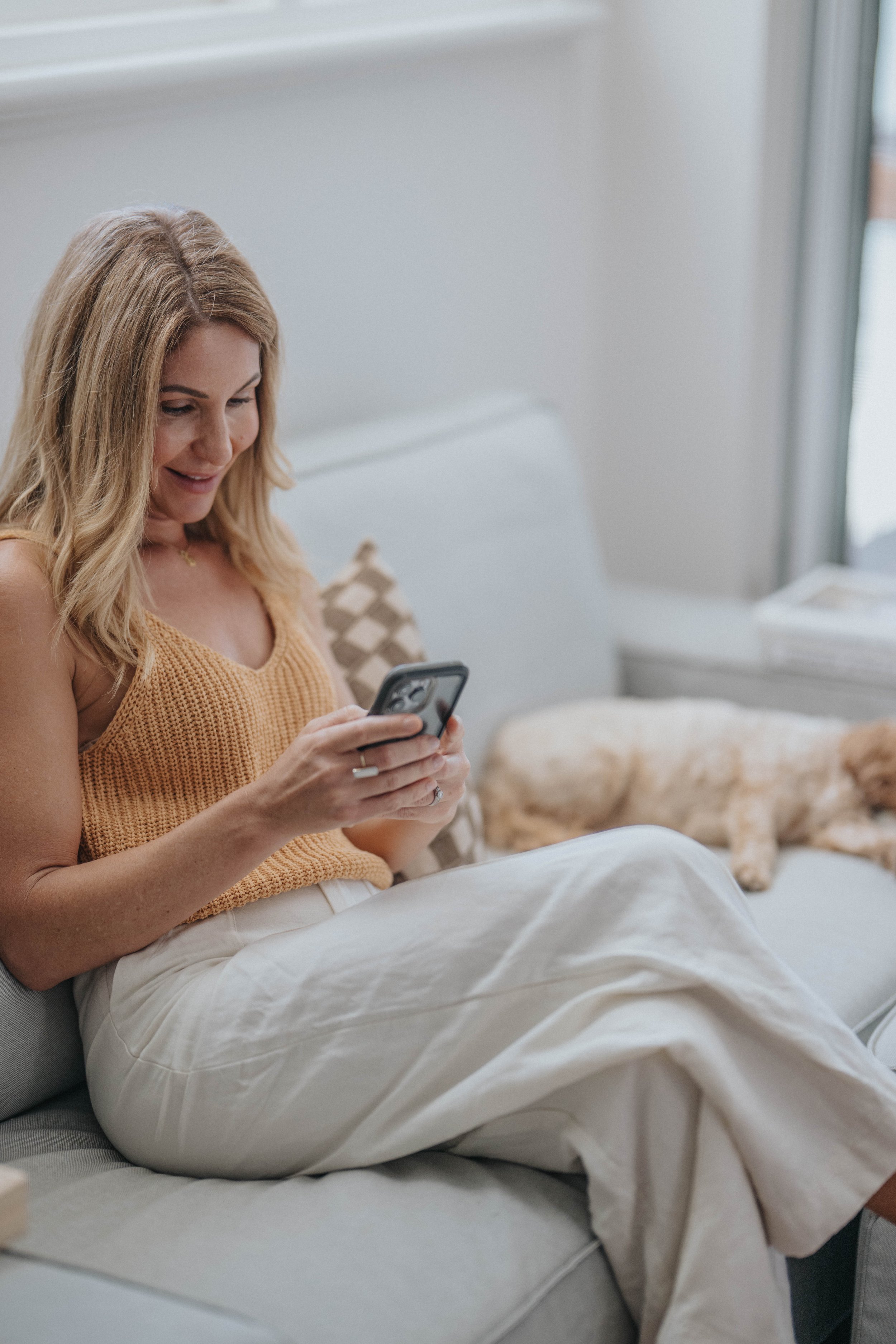 A woman sitting on a light-colored sofa, looking at her phone, with a dog lying beside her.