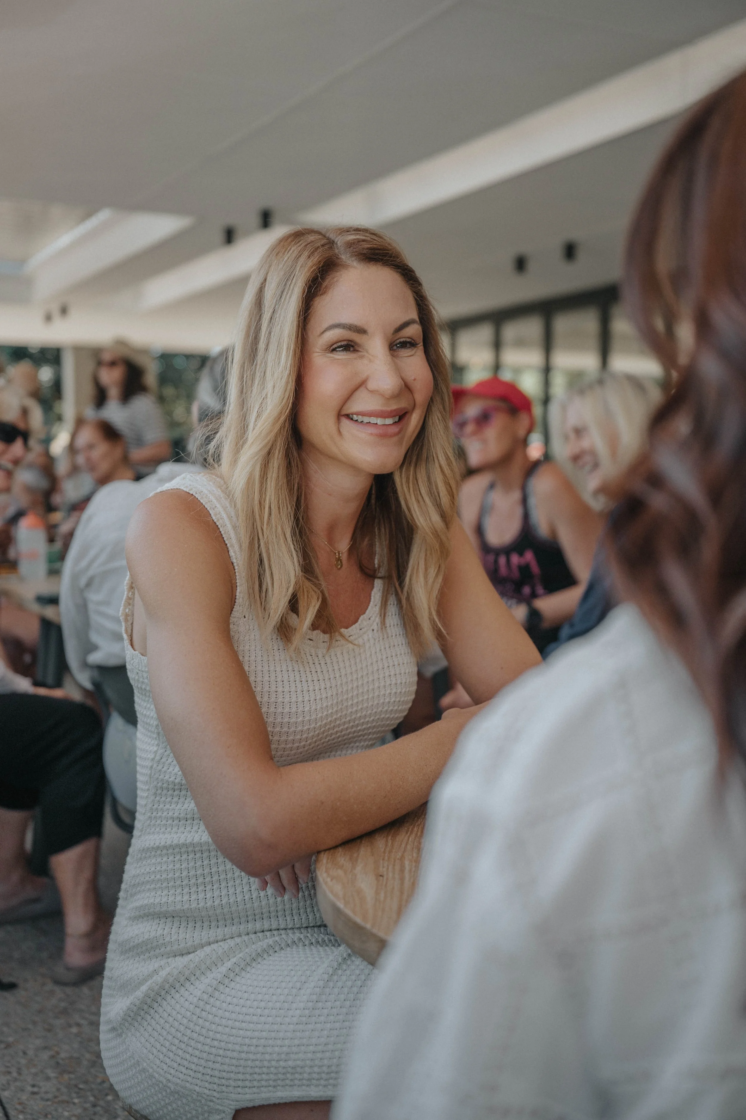 A woman with blonde, wavy hair smiling while talking to another woman in a crowded indoor setting.