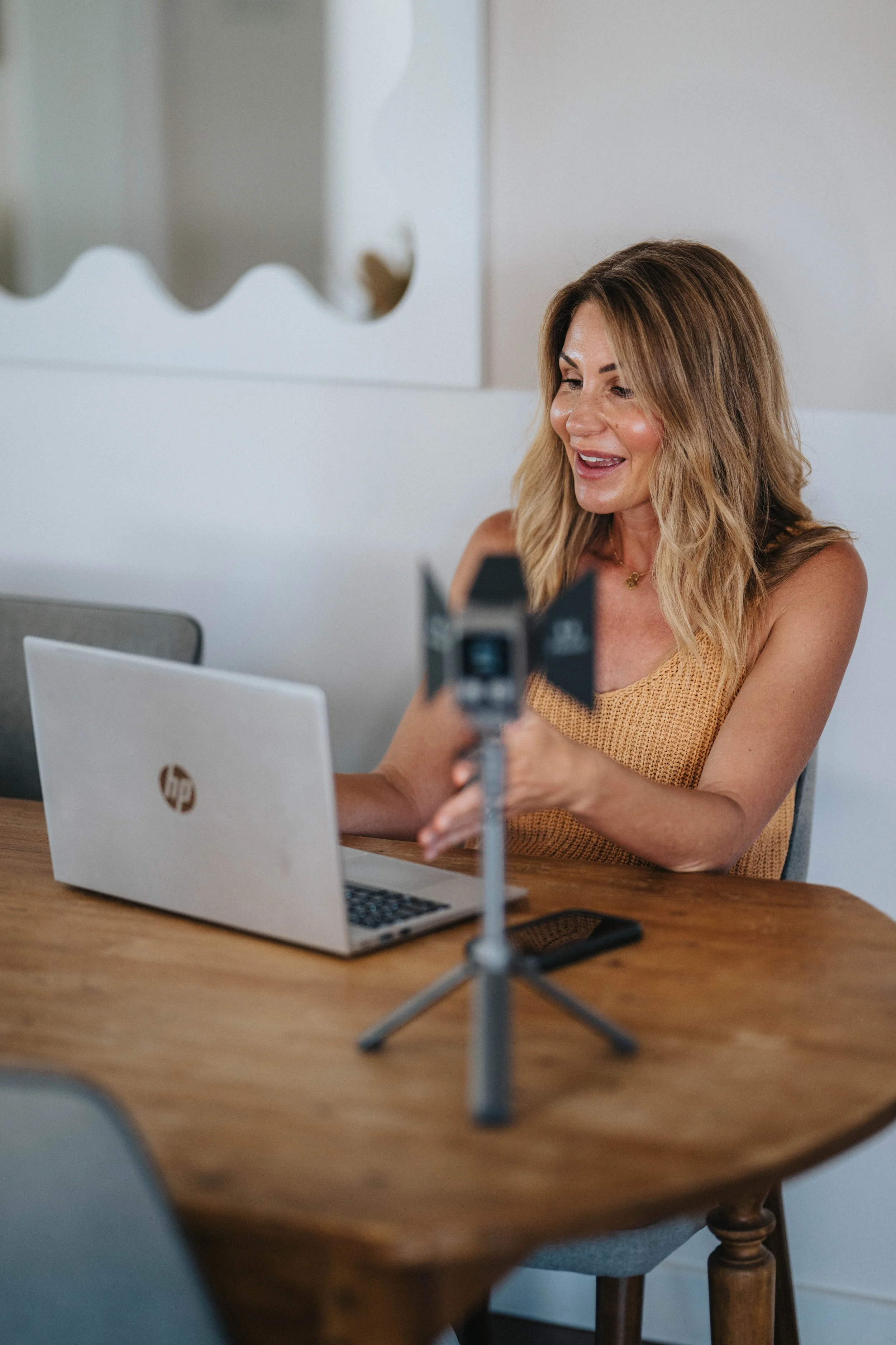 A woman with blonde hair sitting at a wooden table in front of a laptop, recording a video using a ring light and a camera mounted on a tripod, in a bright indoor space.