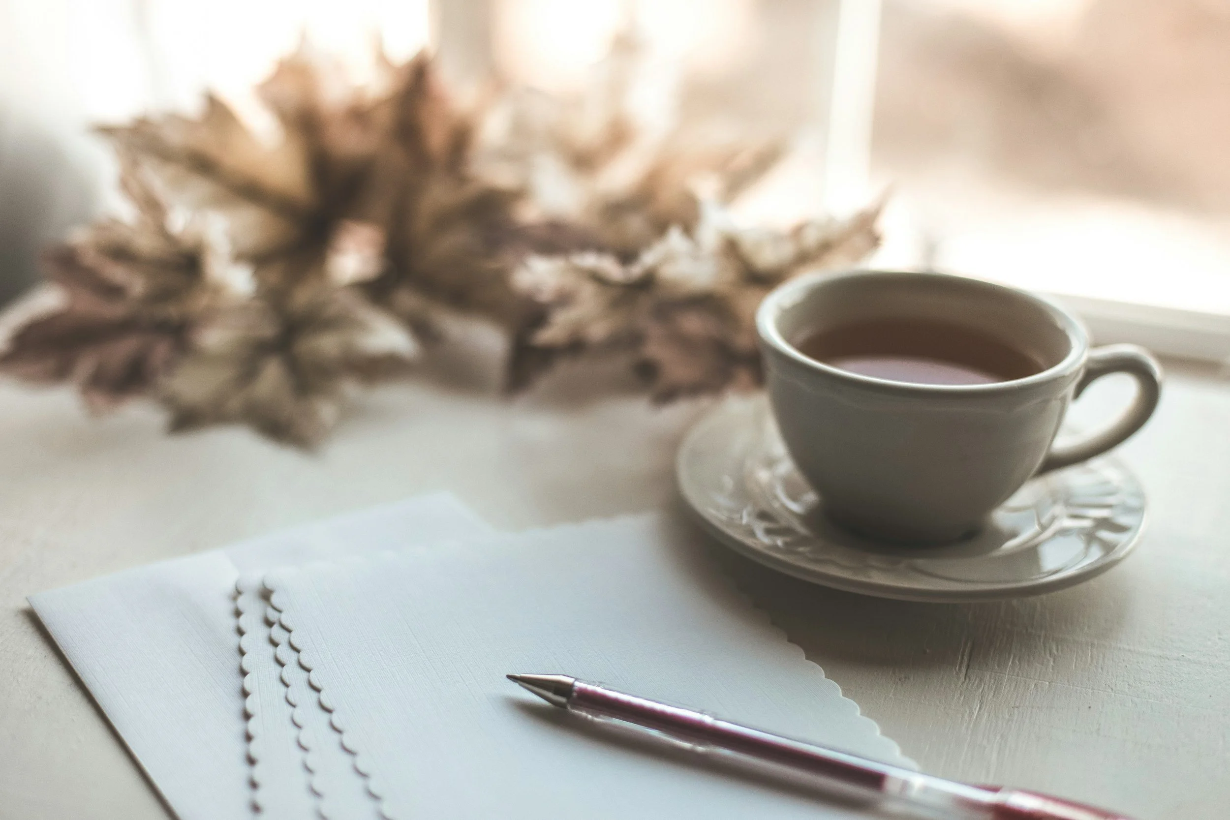 A white cup of tea on a matching saucer placed on a windowsill, with a pen and sheets of decorative paper with scalloped edges in the foreground, and a blurred dried flower arrangement in the background.