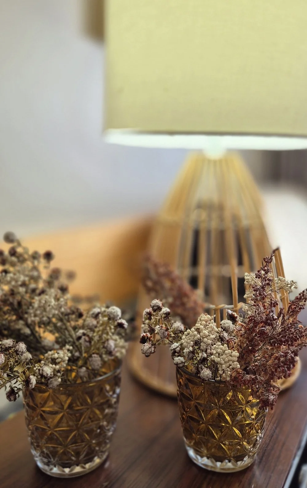Decorative dried flowers in vintage glass jars placed on a wooden surface with a yellow lampshade in the background.