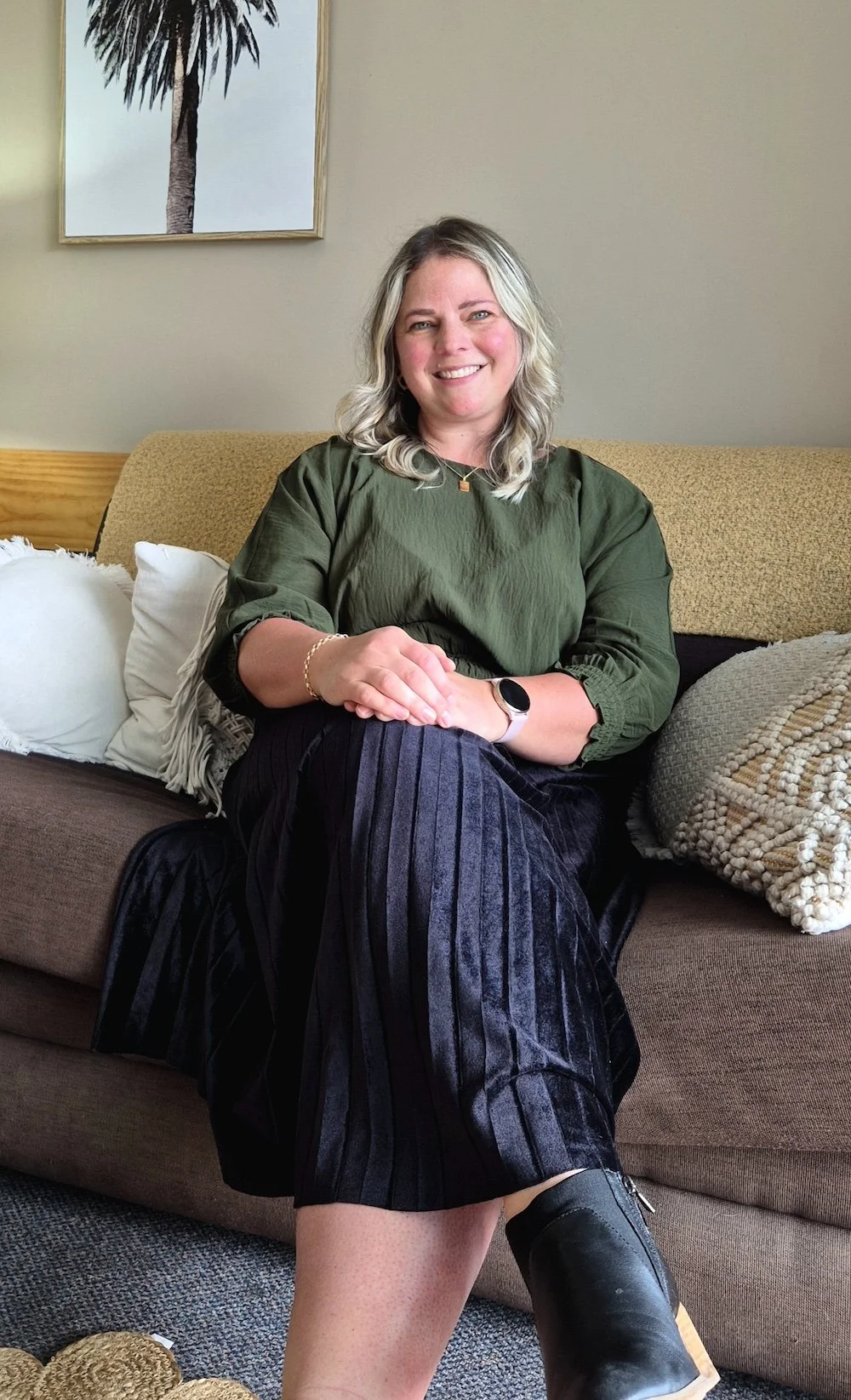 A woman sitting on a sofa, wearing a green top, black pleated skirt, and black boots, smiling at the camera, with a painting of a palm tree on the wall behind her.