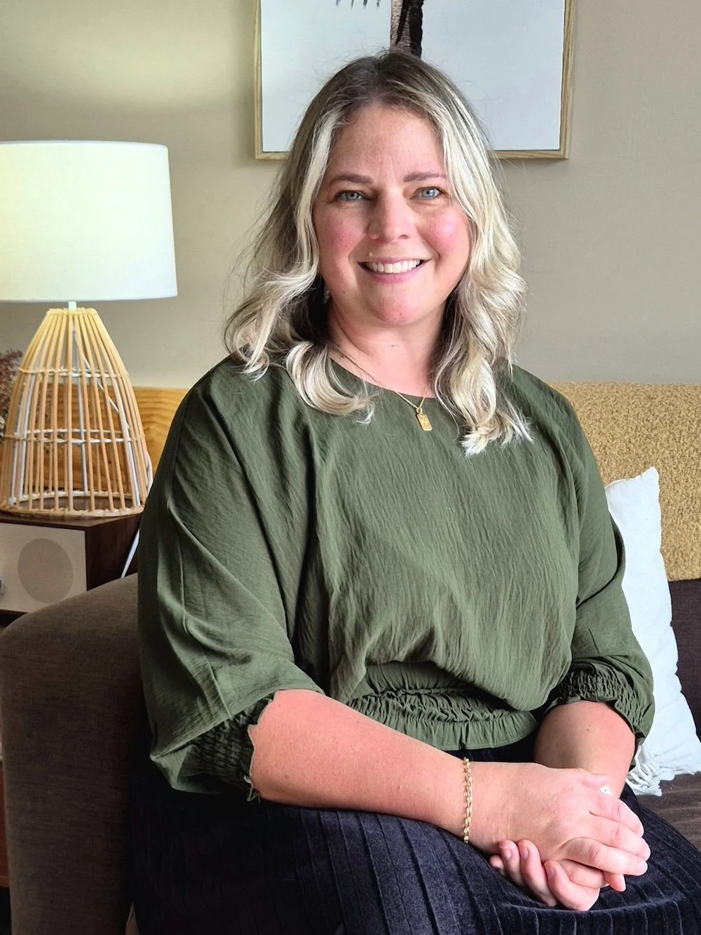 A woman with blonde hair smiling while sitting on a couch in a living room, wearing a green blouse and a gold necklace.