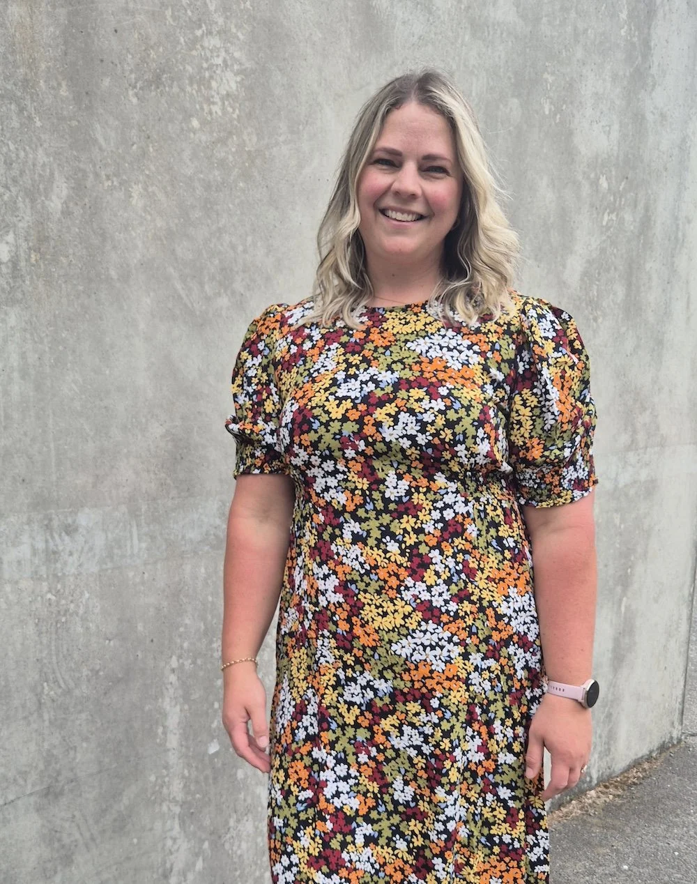 A woman with shoulder-length blonde hair standing and smiling in front of a concrete wall, wearing a colorful floral dress and a smartwatch.