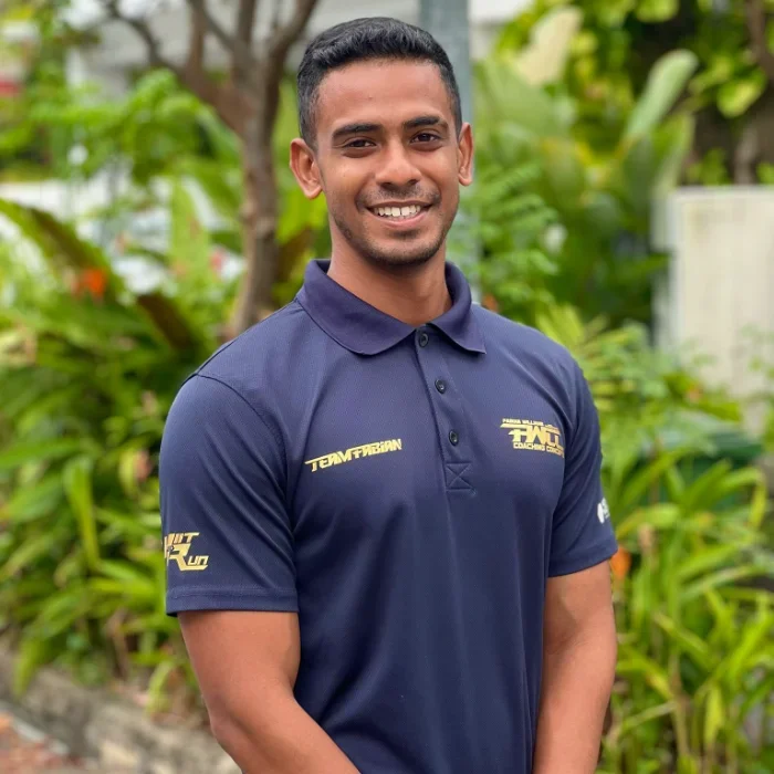 Young man smiling outdoors, wearing a navy blue polo shirt with yellow logos, surrounded by green plants.