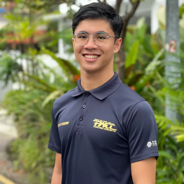A young man with black hair and glasses smiling outdoors, wearing a navy blue polo shirt with yellow and white logos, surrounded by green plants and trees.