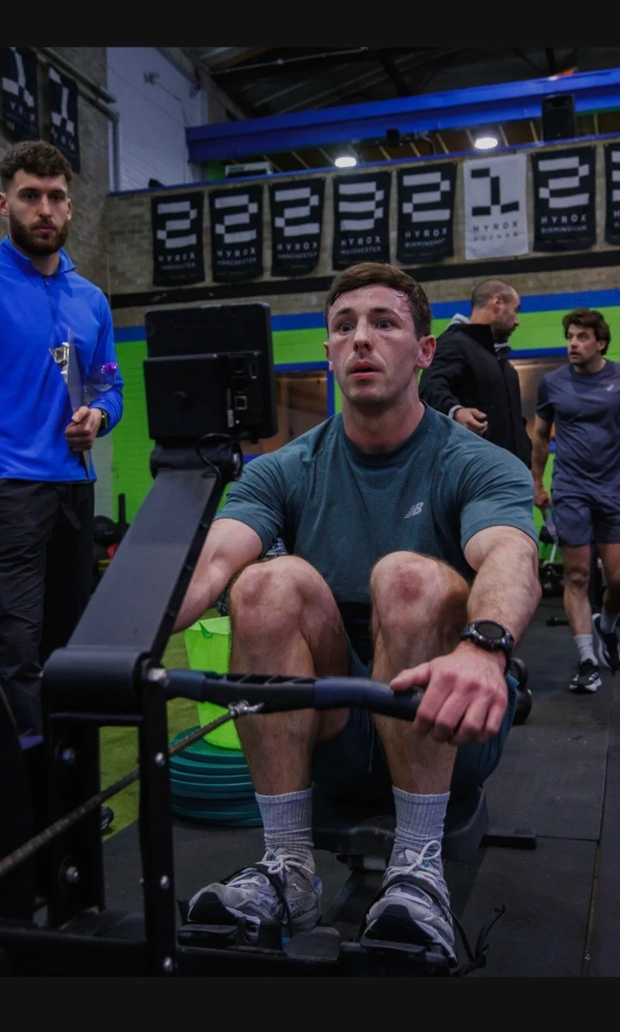 Man using rowing machine at gym, surrounded by other people and gym equipment.