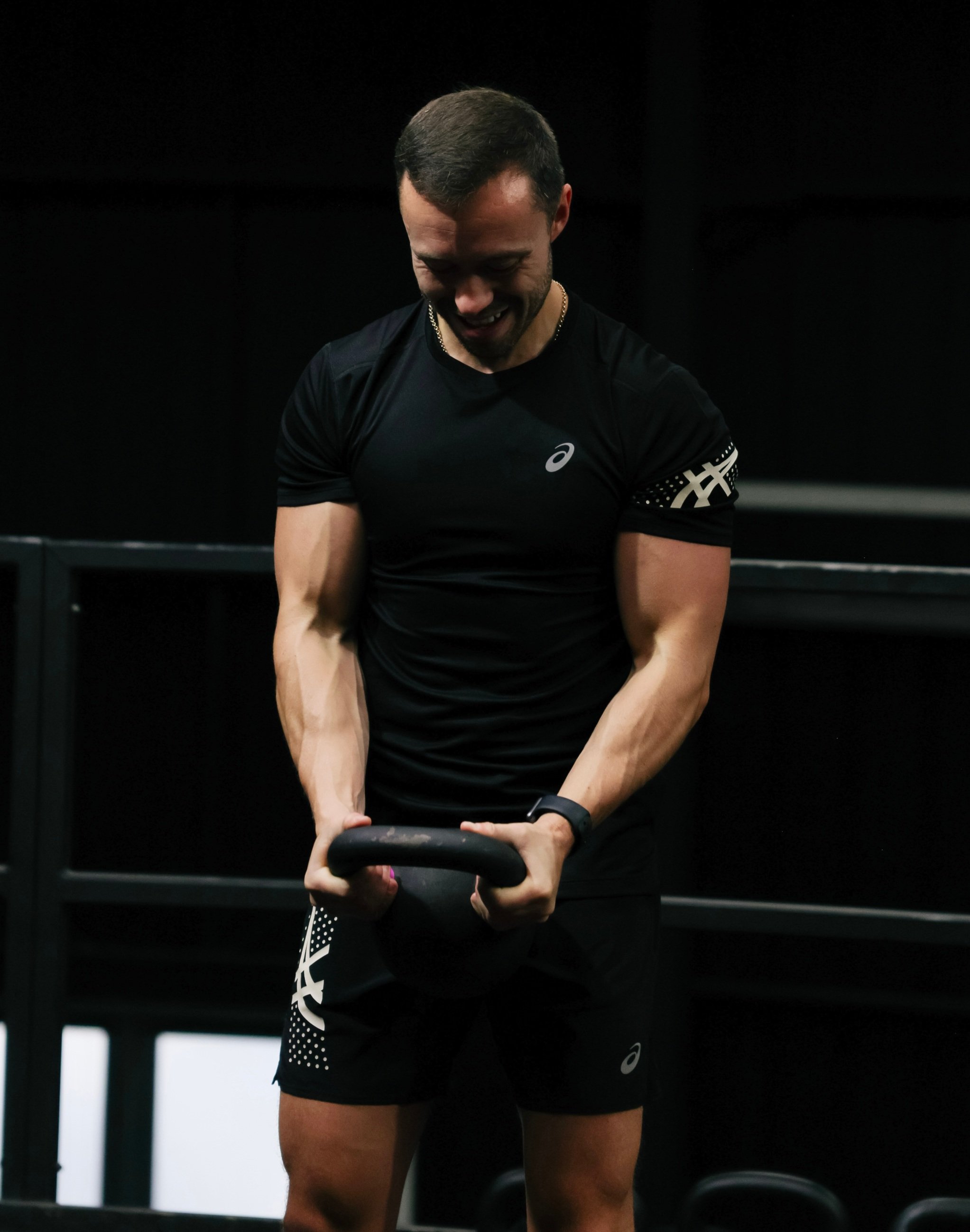 Man in black athletic wear lifting a kettlebell in a dark gym.