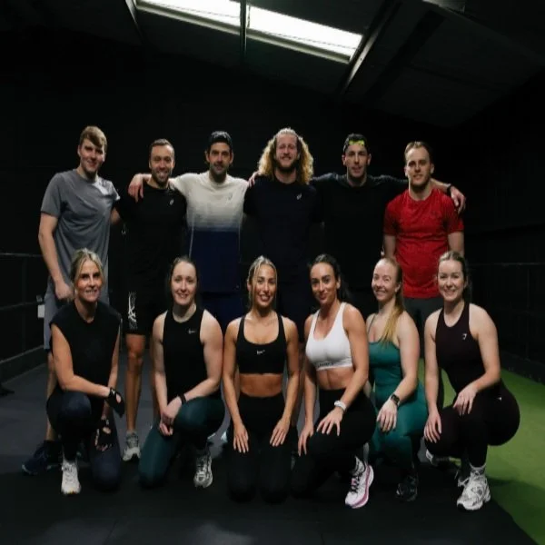 Group photo of Adam Davies Fitness class members in a modern Liverpool gym. Eleven men and women in activewear pose together after a strength and fitness training session, showcasing a supportive community fitness environment.