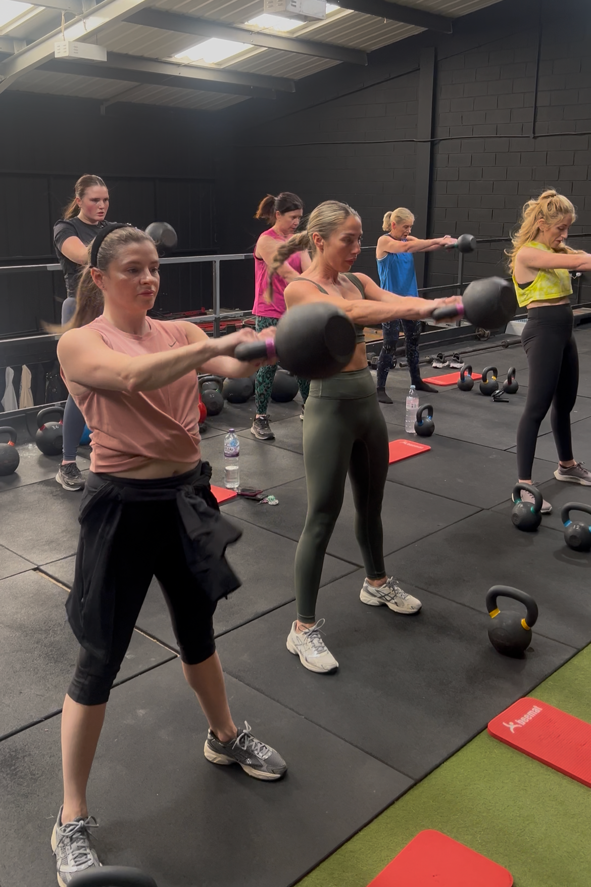 A group of women performing kettlebell swings during a fitness class inside a gym, with kettlebells and exercise mats visible on the floor around them.