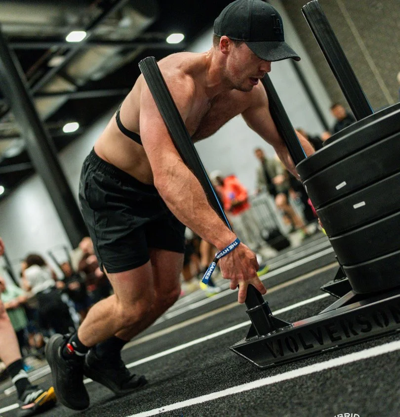 Shirtless man pushing a weighted sled in a gym with text reading “Online Coaching.”