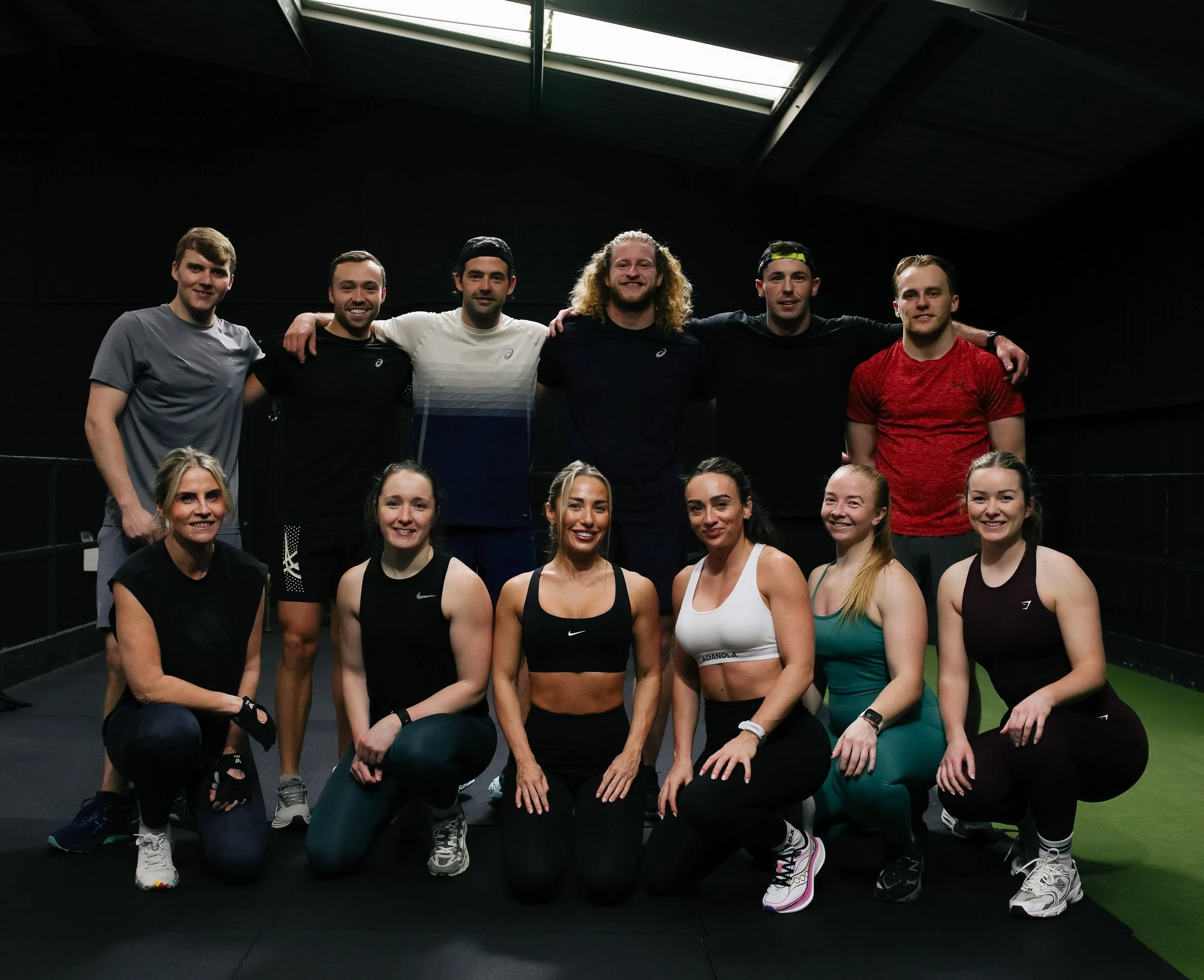 Group photo of Adam Davies Fitness class members in a modern Liverpool gym. Eleven men and women in activewear pose together after a strength and fitness training session, showcasing a supportive community fitness environment.