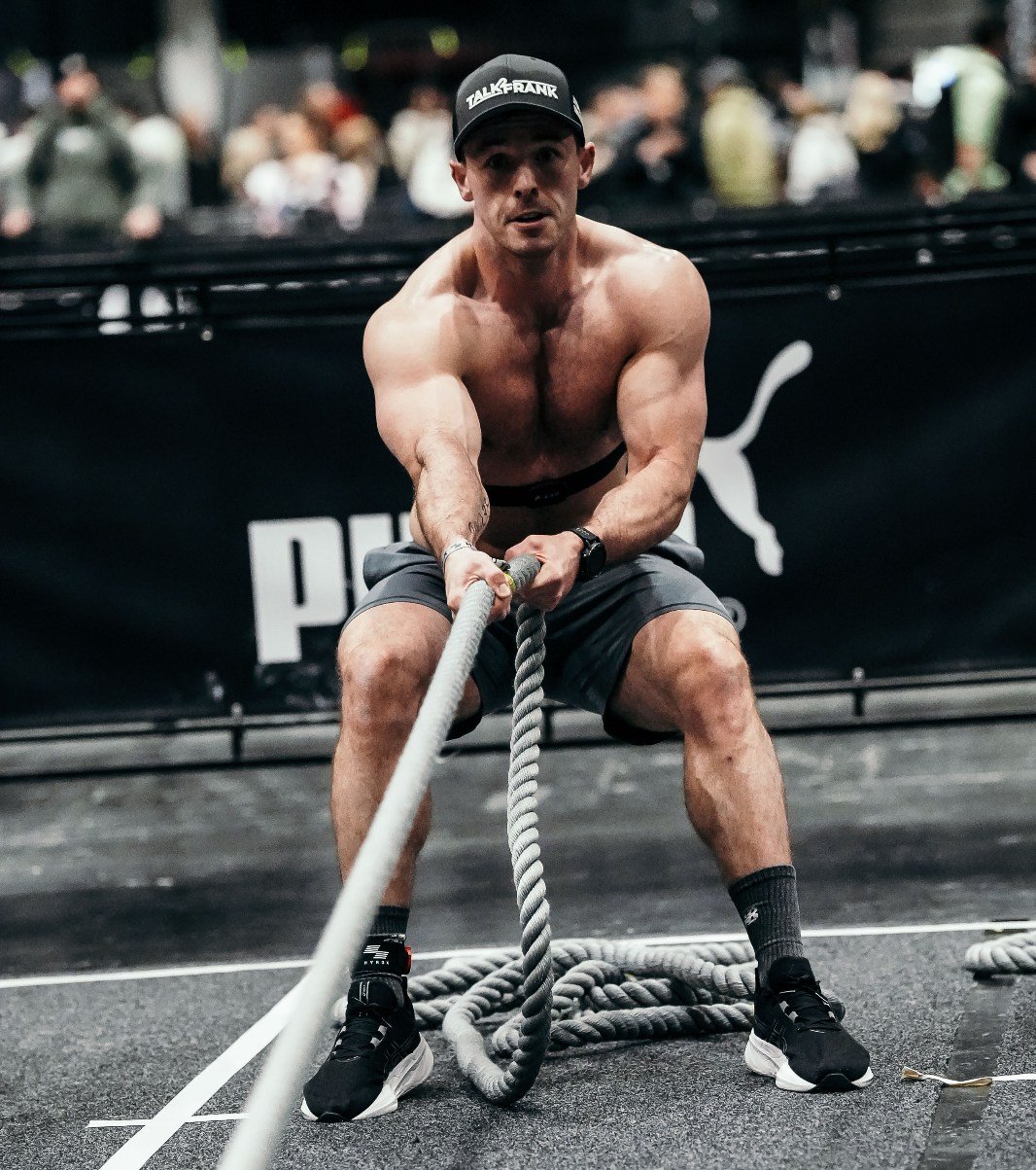 A shirtless man wearing a black cap, black shorts, and black shoes is pulling a thick battle rope during a workout. He is in a gym with a black backdrop featuring a Puma logo and a crowd of people in the background.