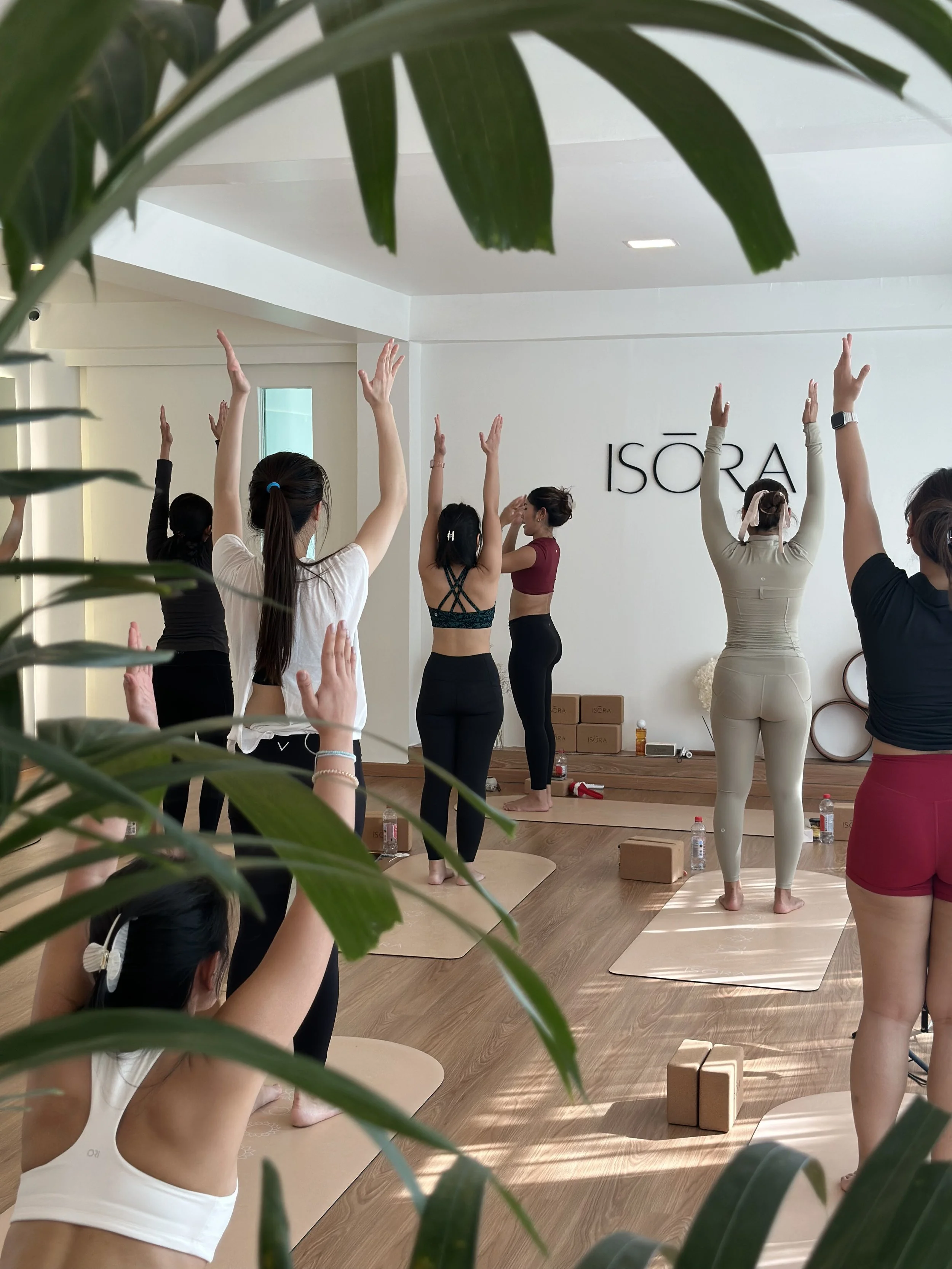Group of people participating in a yoga or fitness class in a studio with the brand name "ISORA" on the wall, some standing with arms raised, with yoga mats and water bottles on the floor.