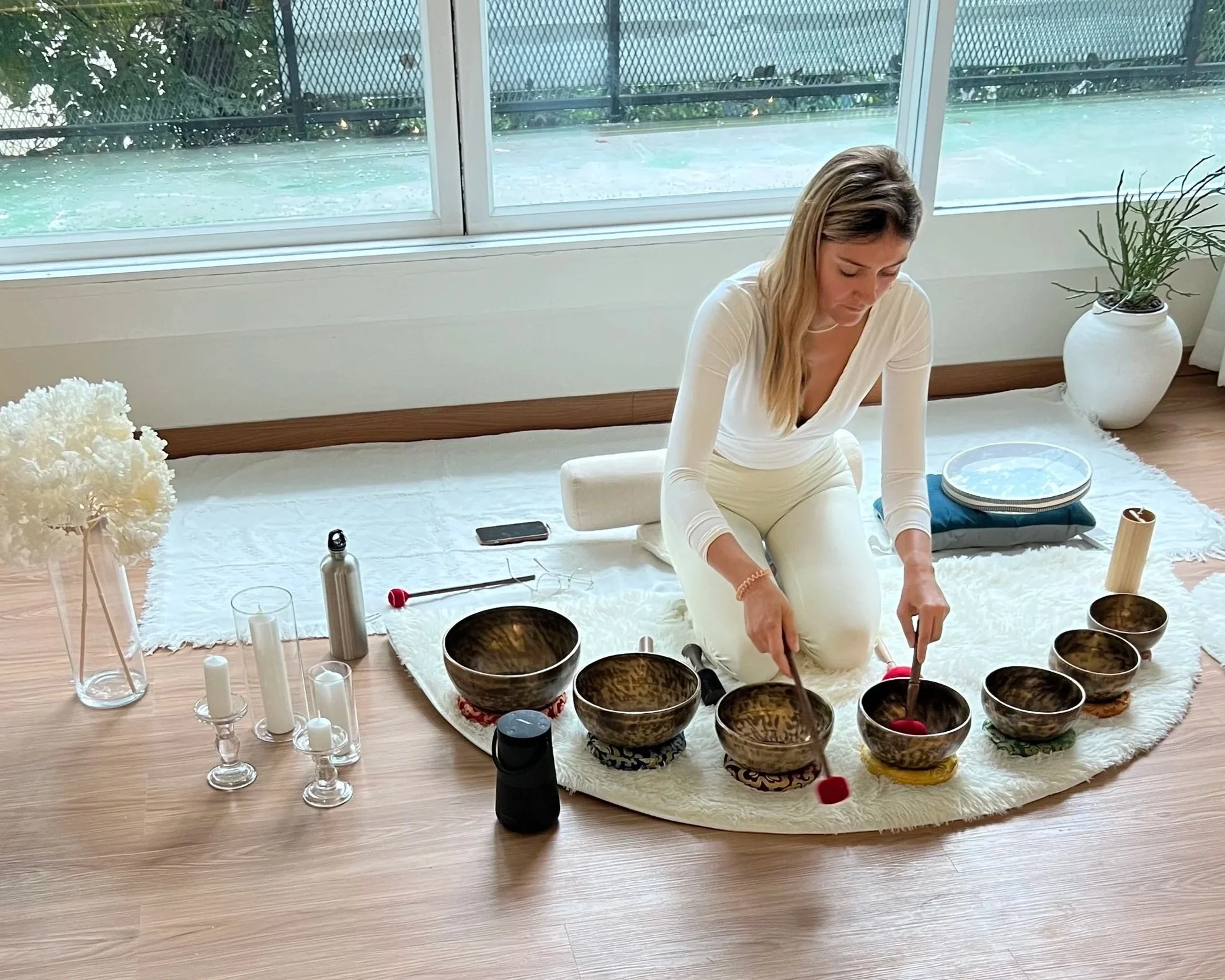 A woman sitting on a white rug inside a room, playing singing bowls with mallets. Glasses, candles, a water bottle, and decorative items are placed around her, with large windows and plants in the background.