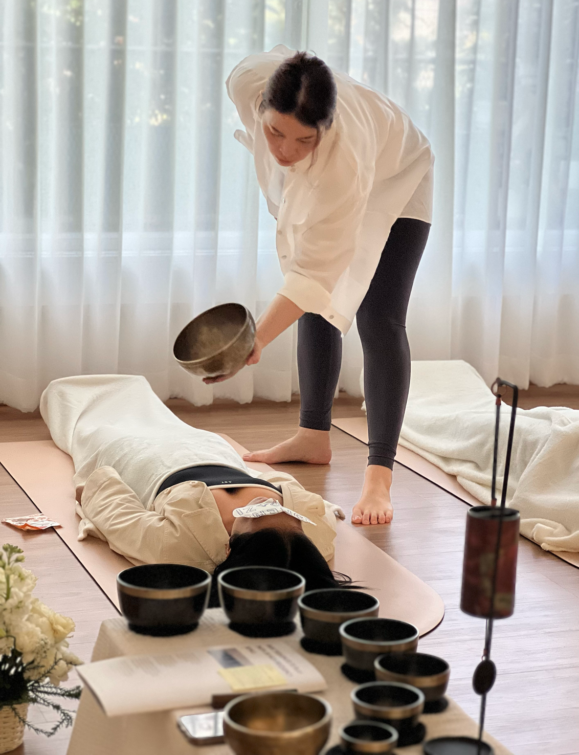 A woman practicing cupping therapy on a woman lying on a yoga mat indoors. The practitioner is holding a cupping glass, and there are multiple cups and therapy items on a table in the foreground, with sheer curtains in the background.