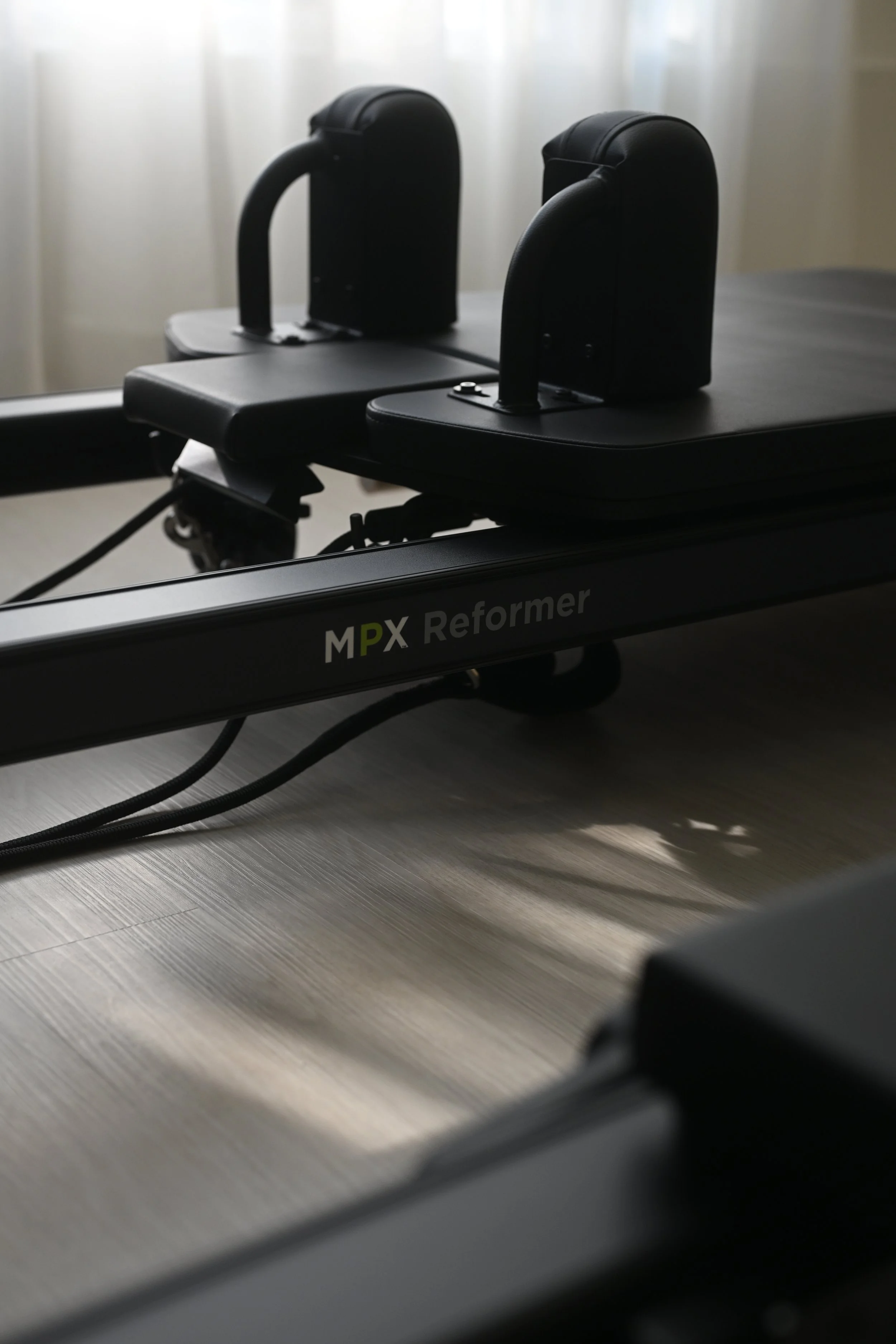 Black MPX Reformer Pilates apparatus on a wooden floor near a window with white curtains.
